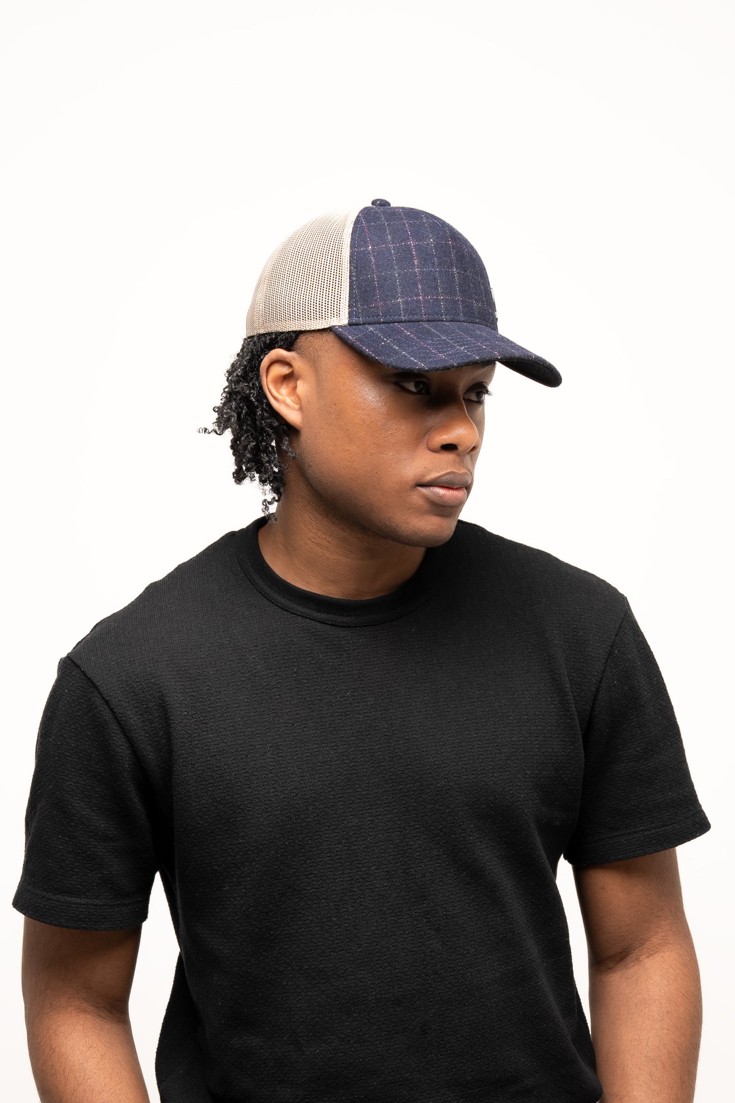 Person wearing a black t-shirt and the Guinness Checked Trucker Cap from Guinness Webstore UK, featuring blue plaid, a beige mesh back, and an adjustable strap, standing against a plain white background.