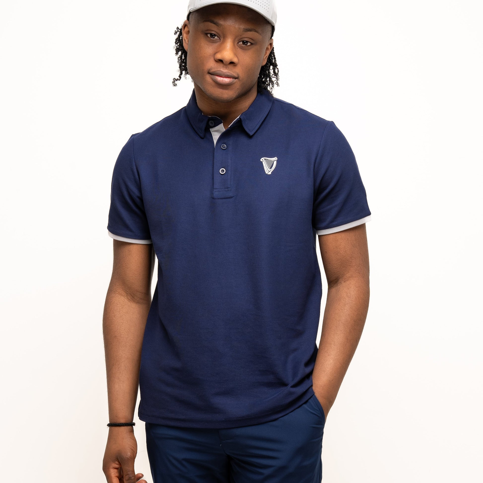 A person wearing a light gray cap, navy blue polo shirt, and Guinness Webstore UK's Guinness Performance Shorts - Navy with a stretch waistband stands against a plain white background, one hand in their pocket.