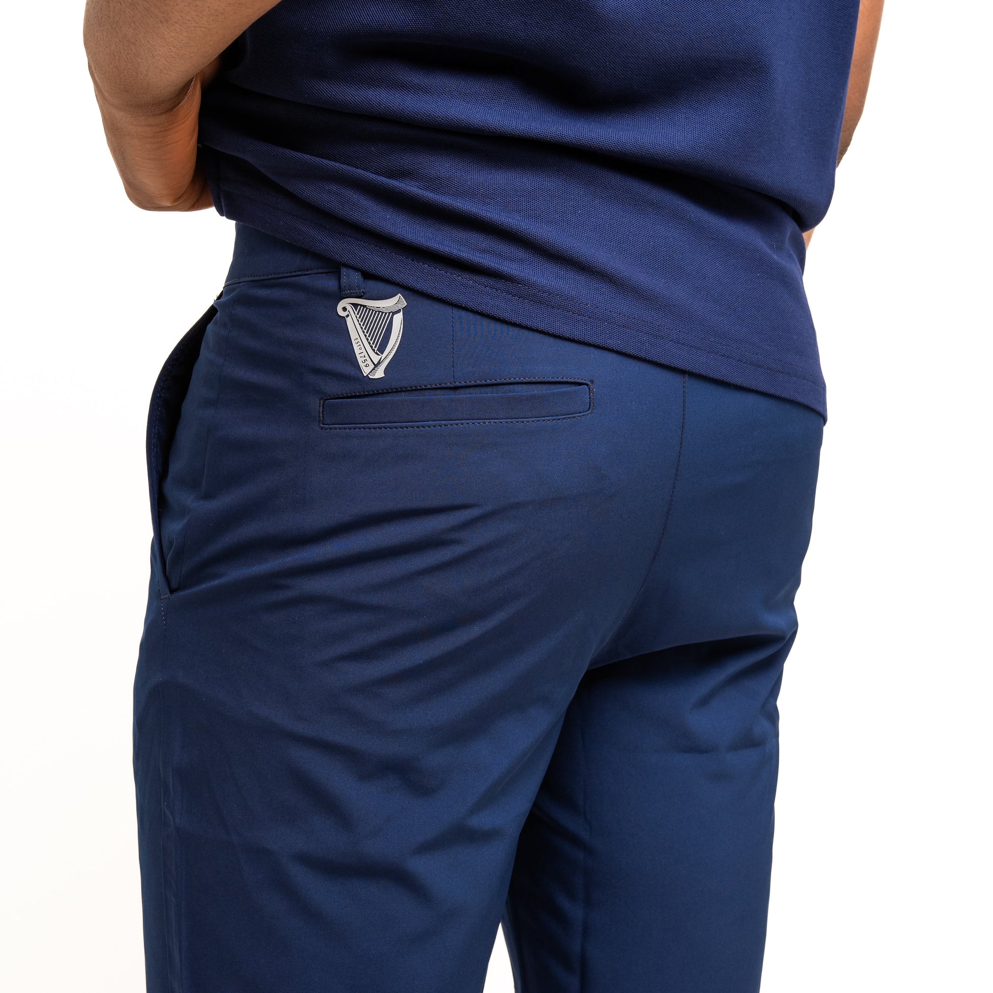 A person wears Guinness Performance Shorts - Navy from Guinness Webstore UK, hands on hips. The shorts have a stretch waistband and a small embroidered logo above the back pocket.