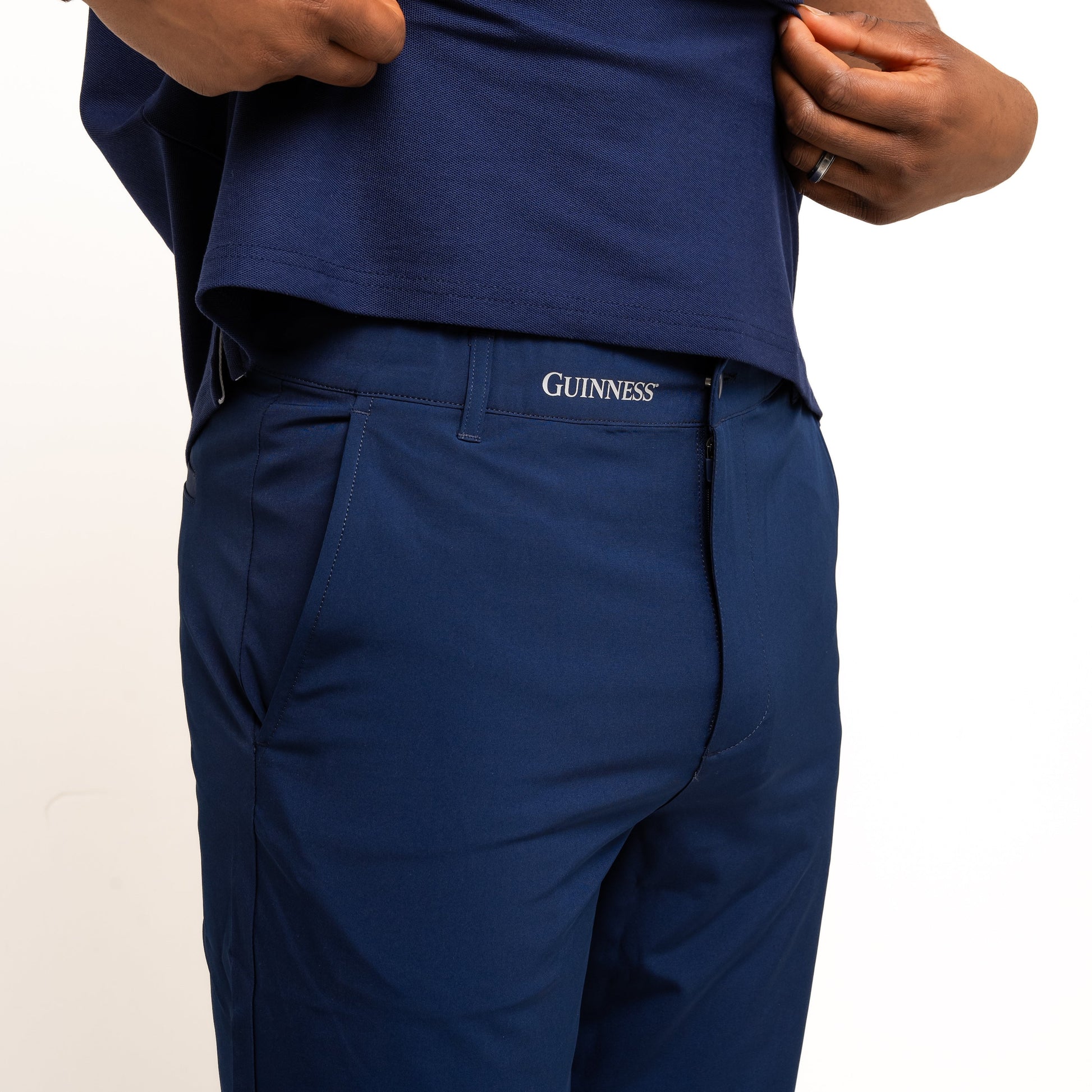 A person wearing a blue polo shirt and navy Guinness Performance Shorts from Guinness Webstore UK, lifting the shirt slightly to reveal the stretch waistband with moisture-wicking fabric for all-day comfort.