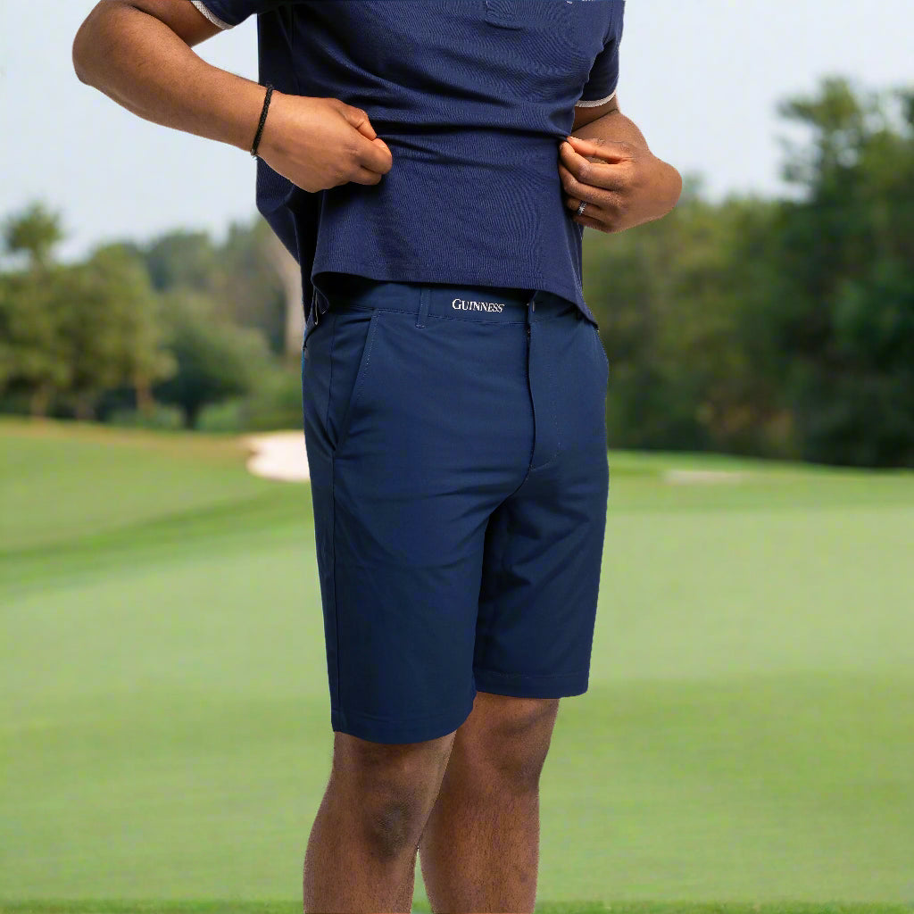 On a golf course with grass and trees in the background, a person wearing a navy blue shirt and Guinness Webstore UK's Guinness Performance Shorts - Navy with a stretch waistband holds up their shirt slightly.