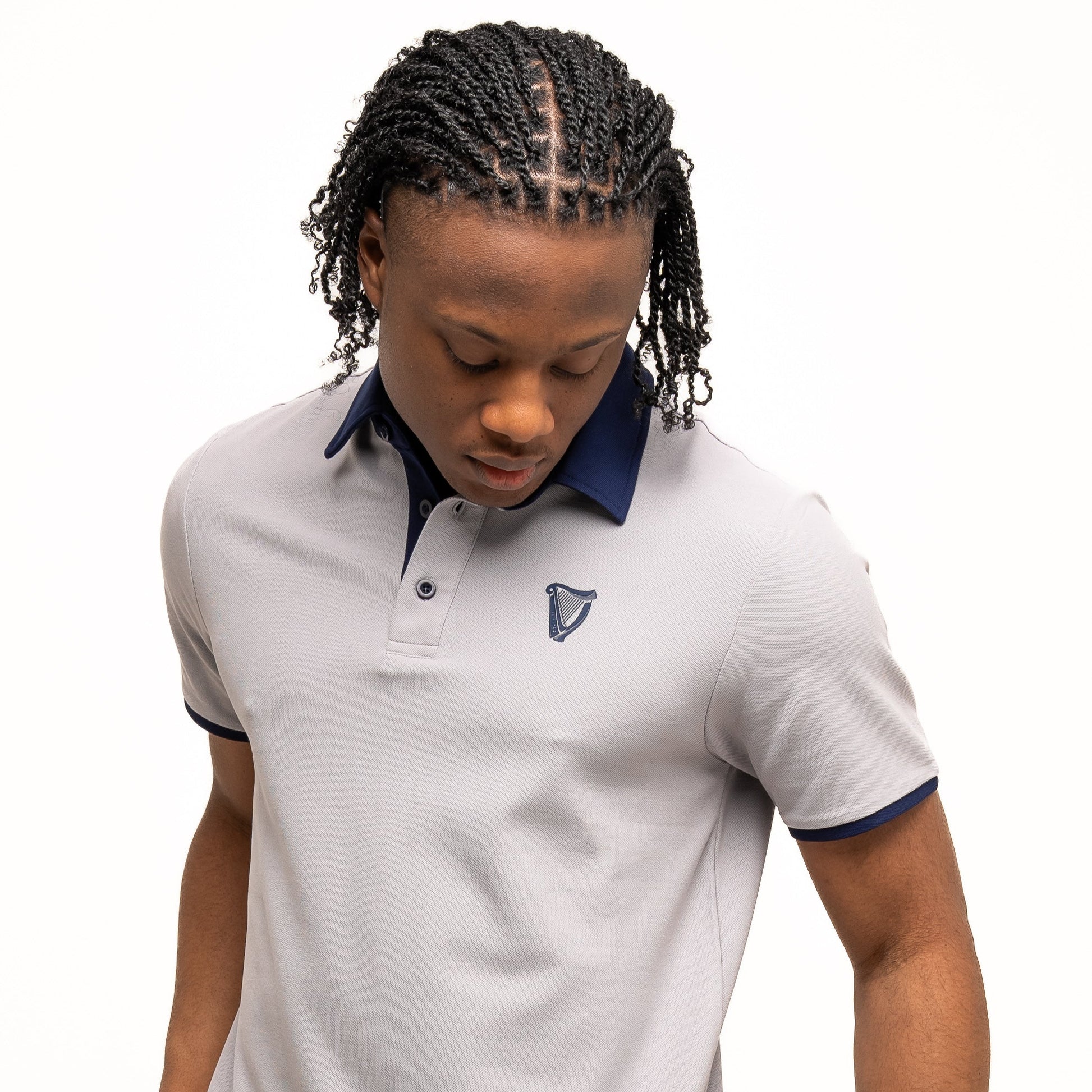 A person with braided hair wears the Guinness Performance Polo - Grey by Guinness Webstore UK, featuring dark blue trim and a small embroidered logo, crafted from premium double pique fabric, looking down while holding the shirt's hem.