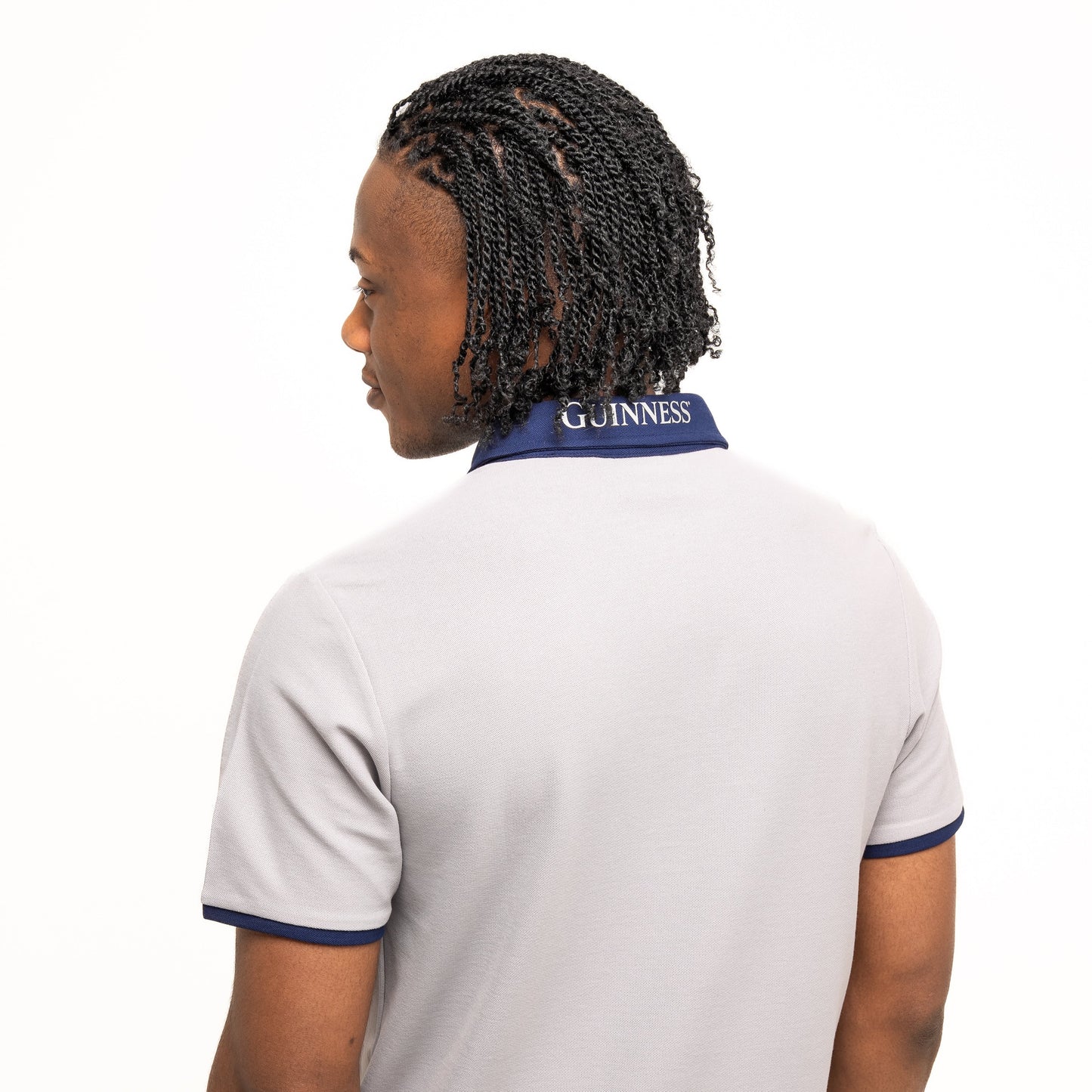 A person stands facing away, wearing the Guinness Performance Polo - Grey from Guinness Webstore UK—a light gray polo with a dark blue collar, "Guinness" text on the back, and made from premium double pique fabric.