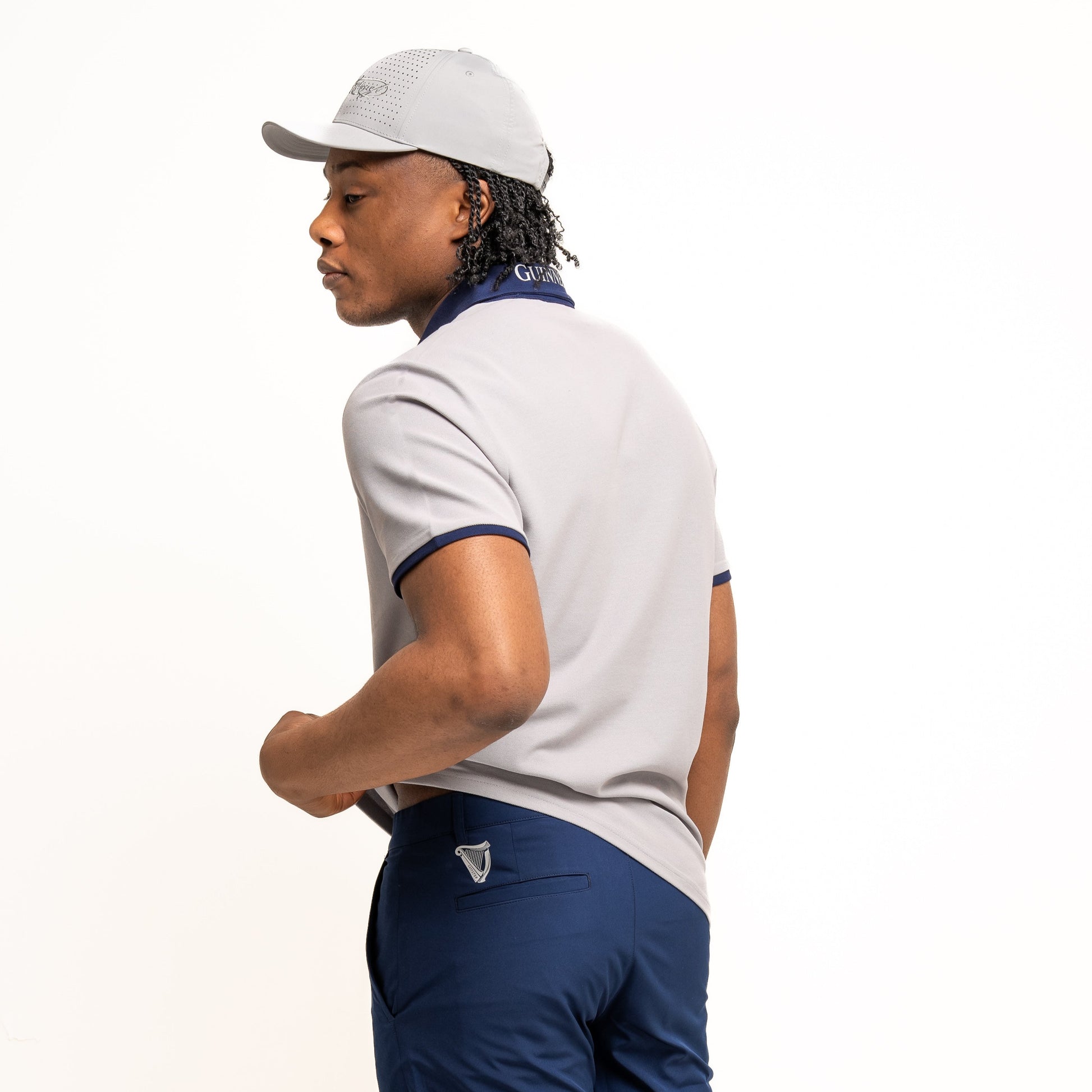 A man in a light gray Guinness Performance Polo by Guinness Webstore UK, navy shorts, and a matching cap stands with his back partly turned to the camera against a white background.