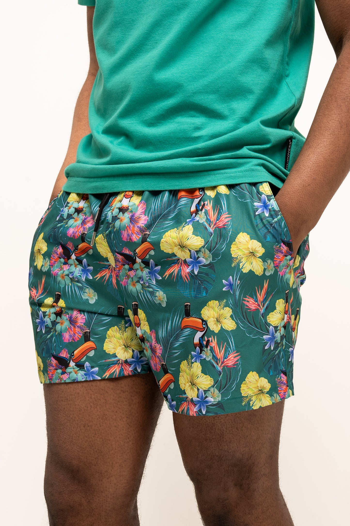 Wearing Guinness UK’s Guinness Toucan Hawaiian Green Beach Shorts and a matching tropical floral tee, someone stands with hands in pockets.