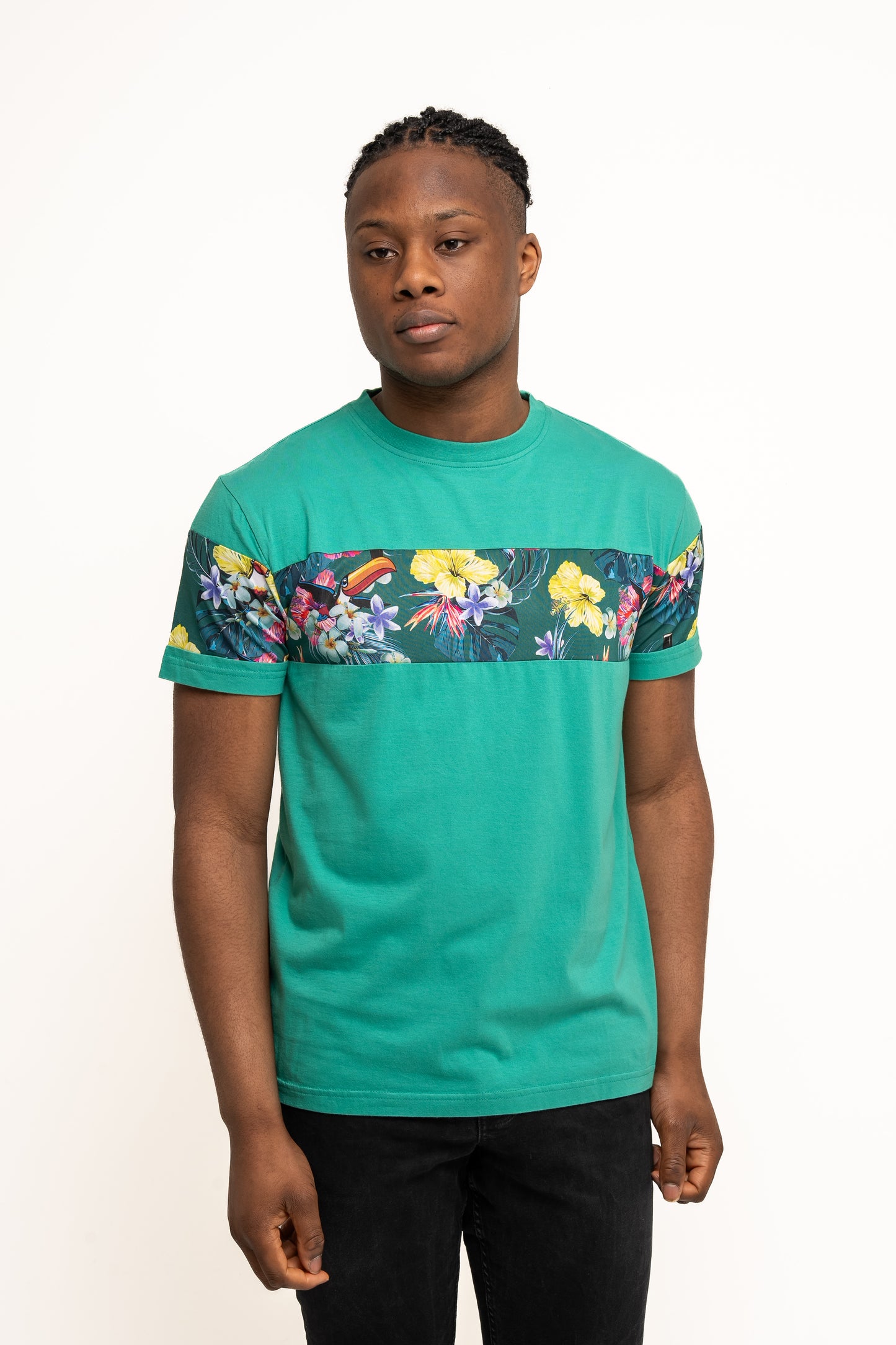 A person stands against a plain background wearing summer wear: the Guinness UK Guinness Toucan Hawaiian Green T-Shirt, featuring a tropical-inspired floral pattern, paired with black pants.