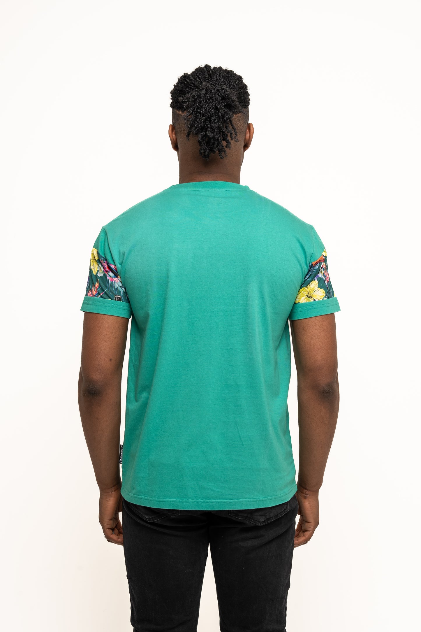 A person with braided hair faces away from the camera, wearing the Guinness UK Guinness Toucan Hawaiian Green T-Shirt with floral-patterned sleeves and black pants—ideal summer attire for a tropical getaway.