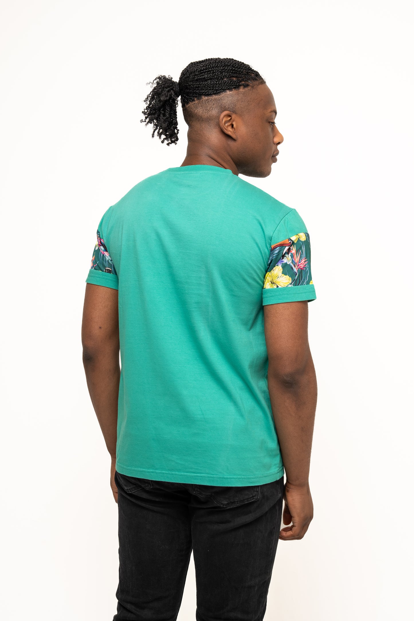 A person faces away, wearing the Guinness Toucan Hawaiian Green T-Shirt by Guinness UK, featuring floral patterned sleeves and paired with black jeans against a plain white background.