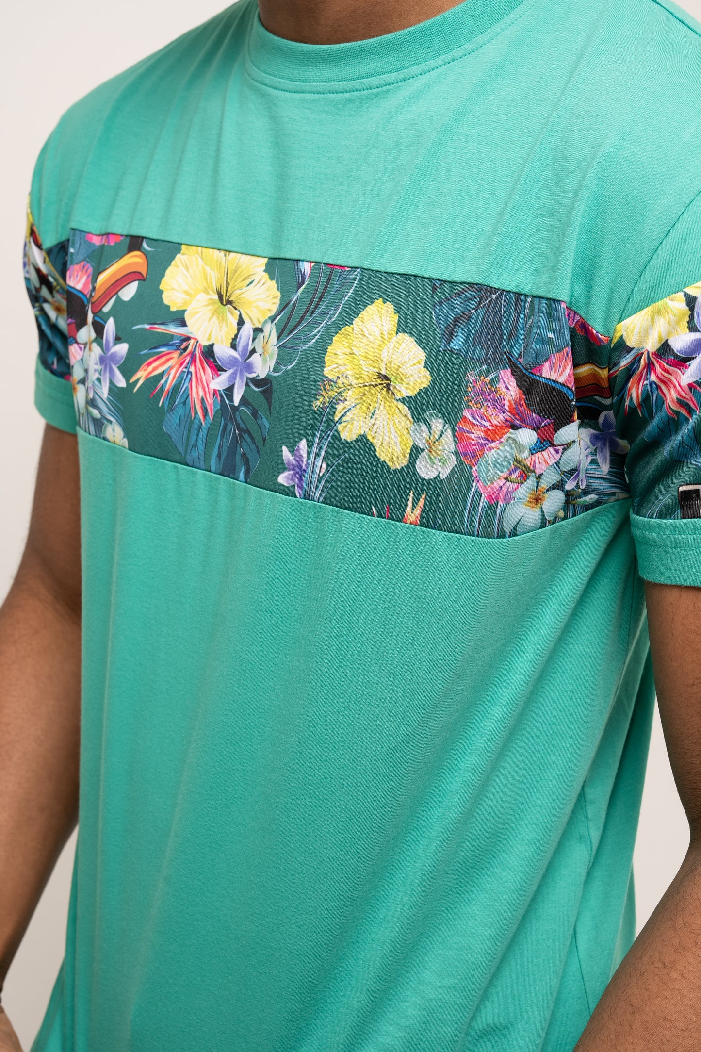 A person wears the Guinness Toucan Hawaiian Green T-Shirt by Guinness UK, featuring a horizontal floral and bird print across the chest and sleeves, capturing a tropical vibe.