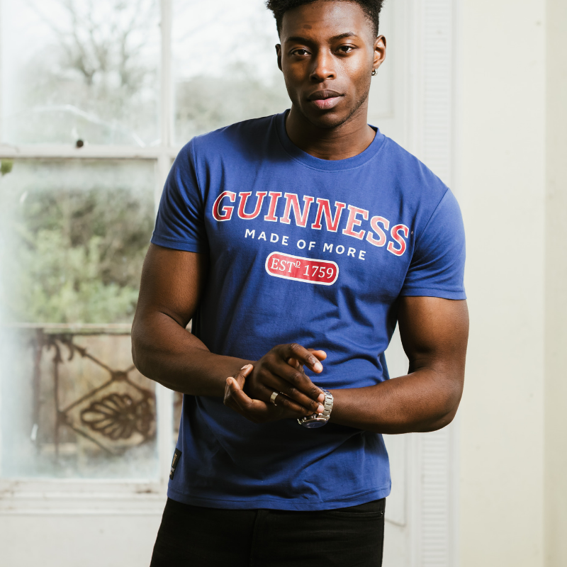 A man wearing a cotton Guinness Blue Trademark Label T-Shirt standing in front of a window.
