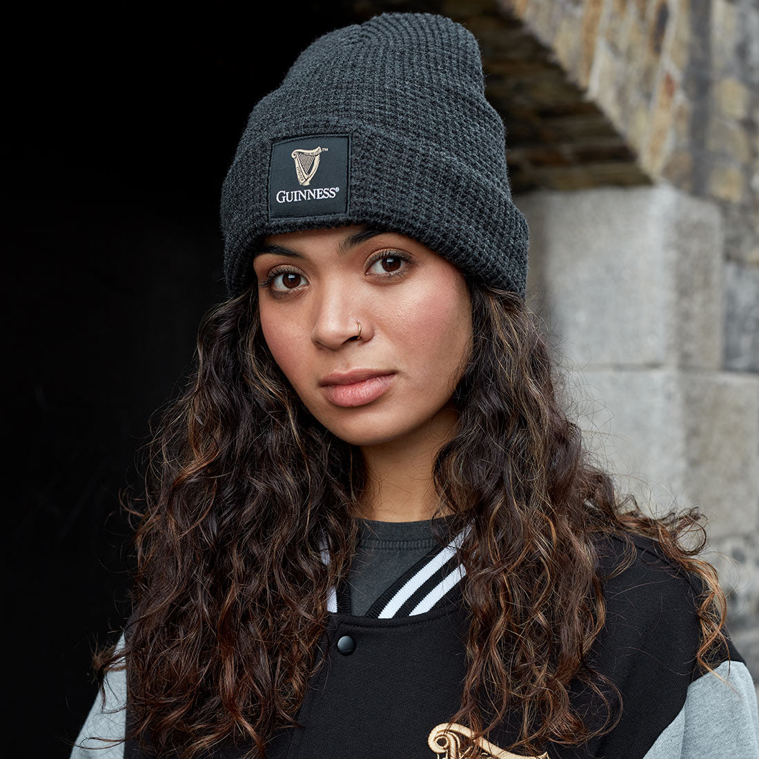 A person with long curly hair wears a black Guinness Thermal Warm Beanie by Guinness UK and a black jacket, standing in front of a stone wall and looking at the camera.