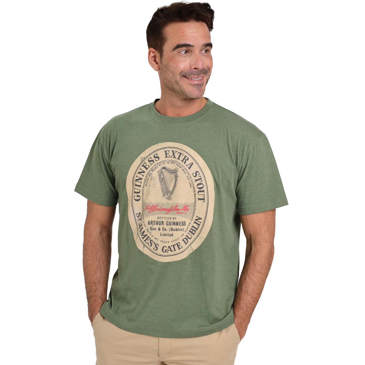 A man in a Guinness UK Guinness Extra Stout Label Green T-Shirt and beige pants stands with his hands in his pockets, smiling slightly, against a plain background. The high-quality poly-cotton fiber and inclusive size range make this shirt a comfortable choice for everyone.