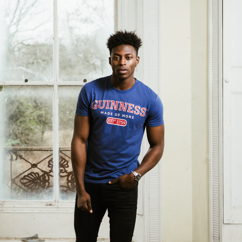 A man wearing a Guinness Blue Trademark Label T-Shirt, by Guinness UK, standing in front of a window.