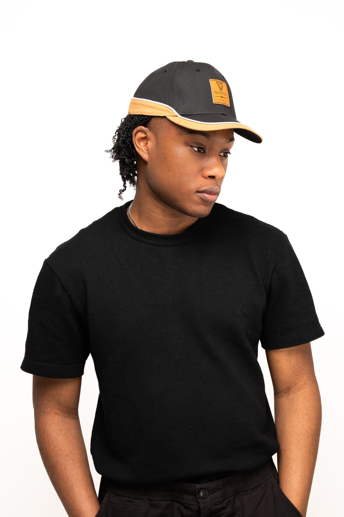 A person wearing a black t-shirt and a Guinness Webstore UK Guinness Black Cap with Caramel Edge looks to their left against a plain white background.
