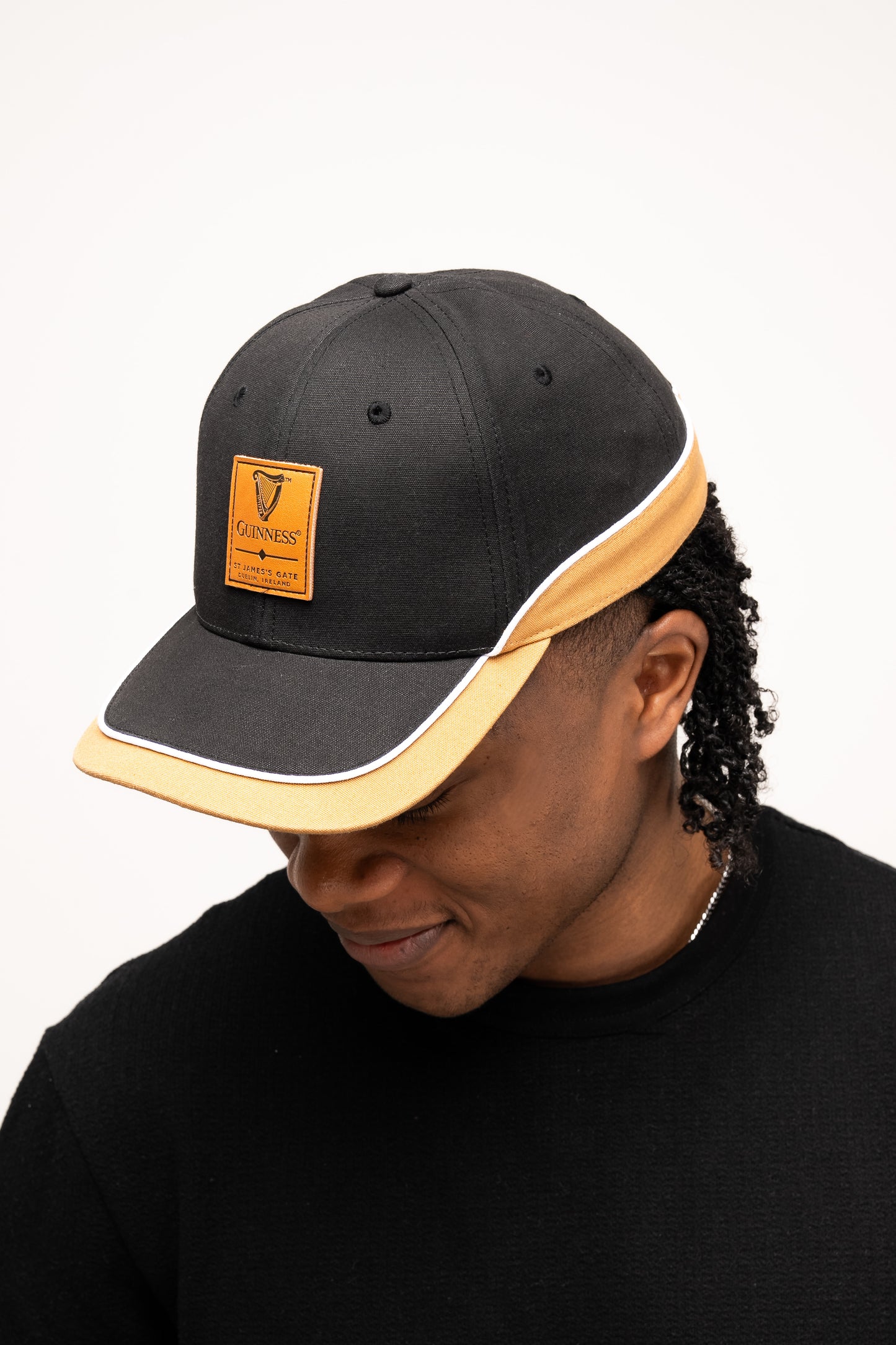 A person wearing a Guinness Webstore UK Black Cap with Caramel Edge looks down, short curly hair peeking out beneath the hat.