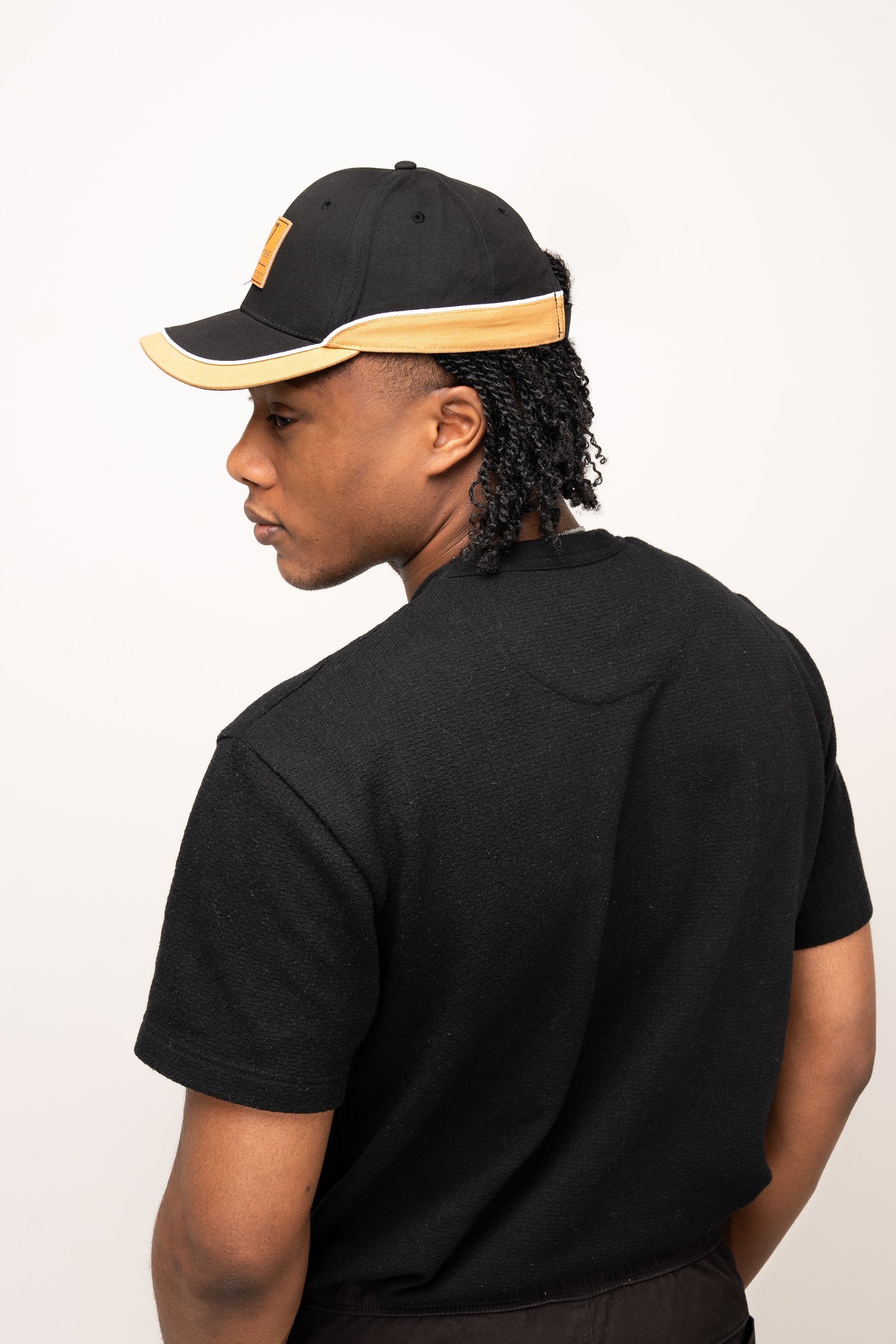 A man in a black t-shirt and the Guinness Black Cap with Caramel Edge from Guinness Webstore UK stands facing away, looking over his left shoulder against a plain background.