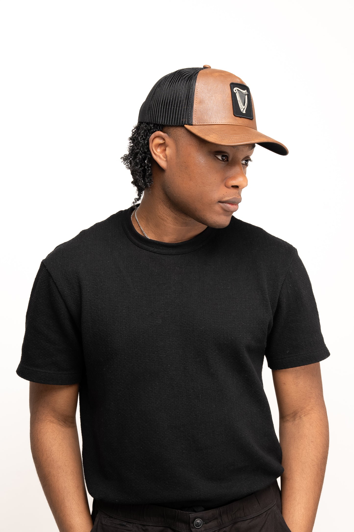 Person wearing a black short-sleeve shirt and a Guinness Brown Leather Trucker Cap from Guinness Webstore UK, featuring the iconic harp logo, standing against a plain white background and looking to the side.