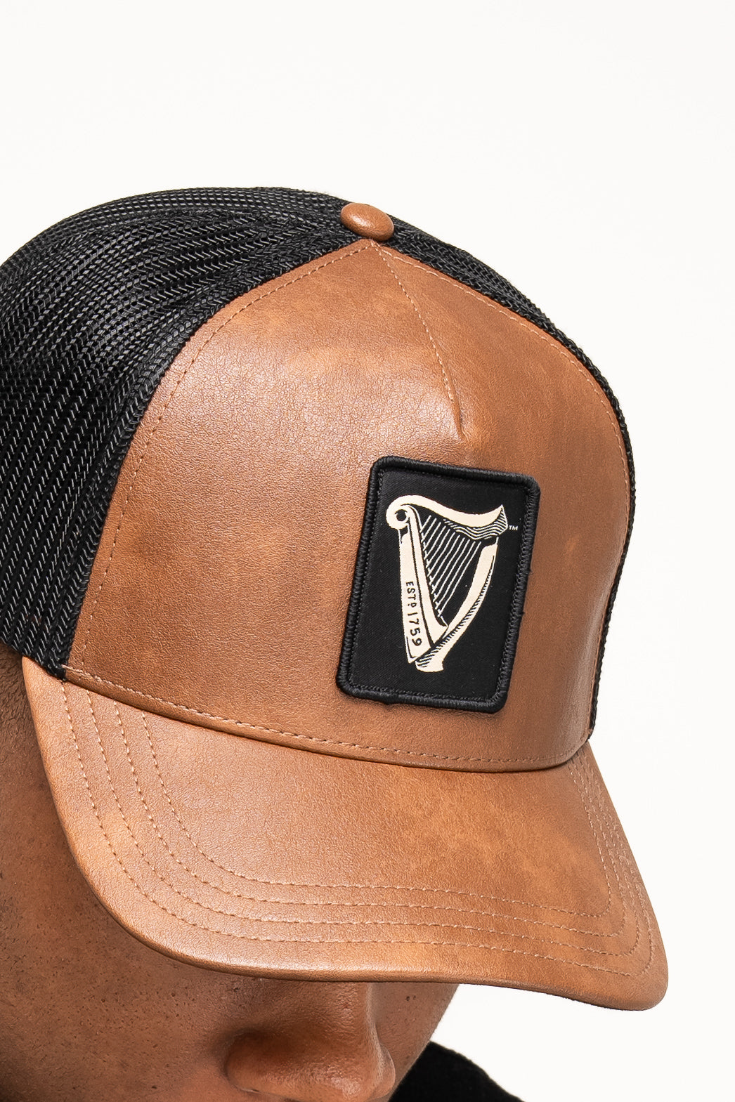 A close-up of the Guinness Brown Leather Trucker Cap from Guinness Webstore UK shows a front panel patch with a harp emblem labeled "Éire," inspired by the classic Guinness harp logo.