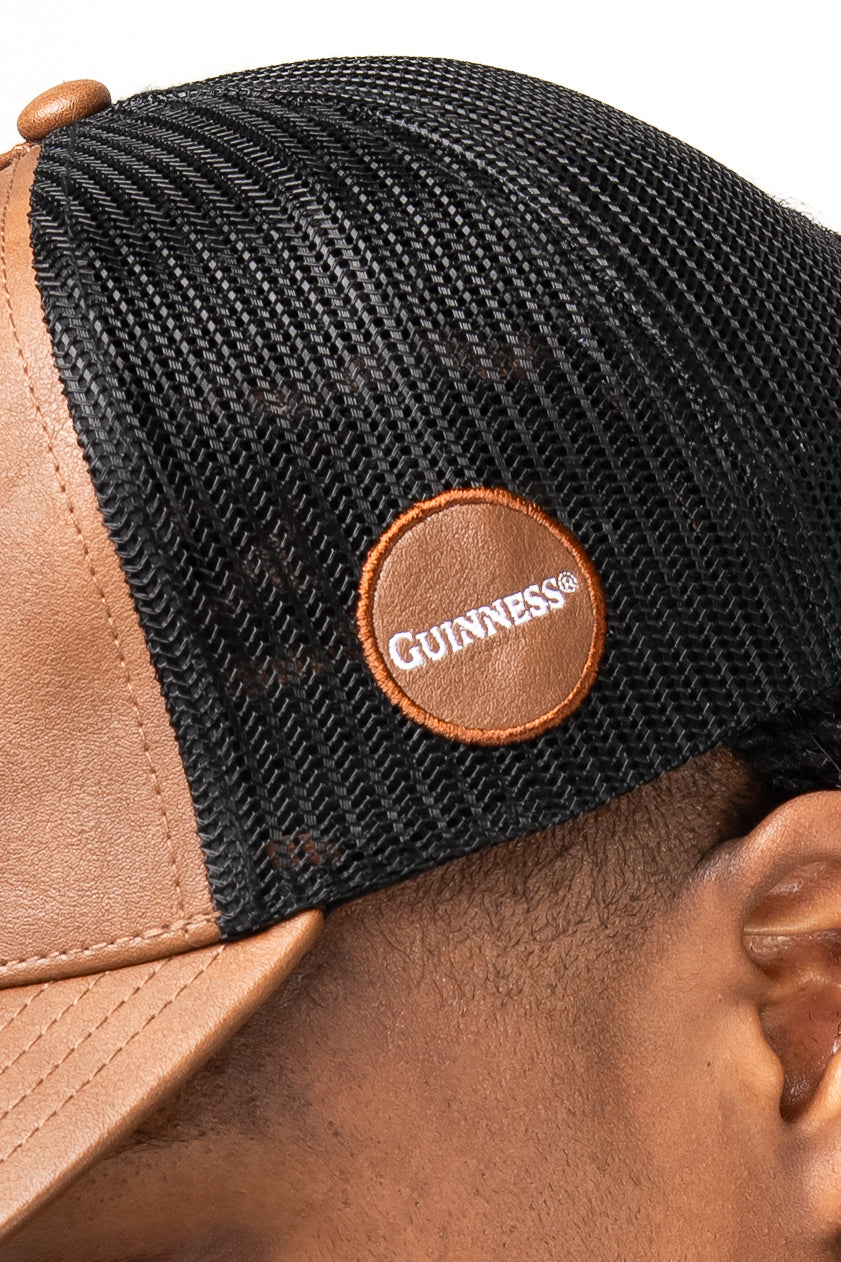 Close-up of a person wearing the Guinness Brown Leather Trucker Cap from Guinness Webstore UK, featuring mesh panels and a round patch with the iconic Guinness harp logo.
