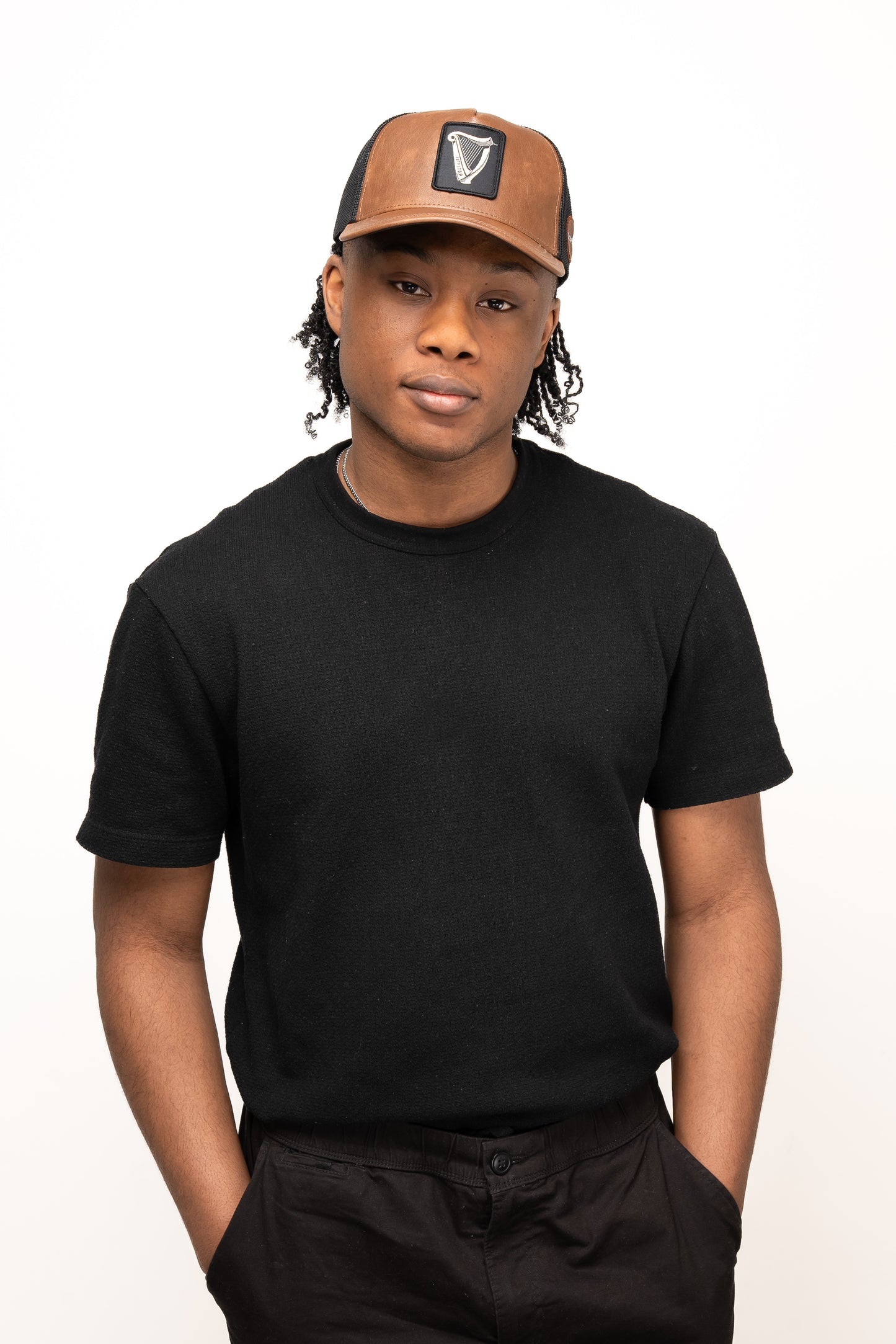 A person in a black t-shirt wears the Guinness Brown Leather Trucker Cap from Guinness Webstore UK, featuring a leather peak and iconic harp logo, standing with hands in pockets against a plain white background.