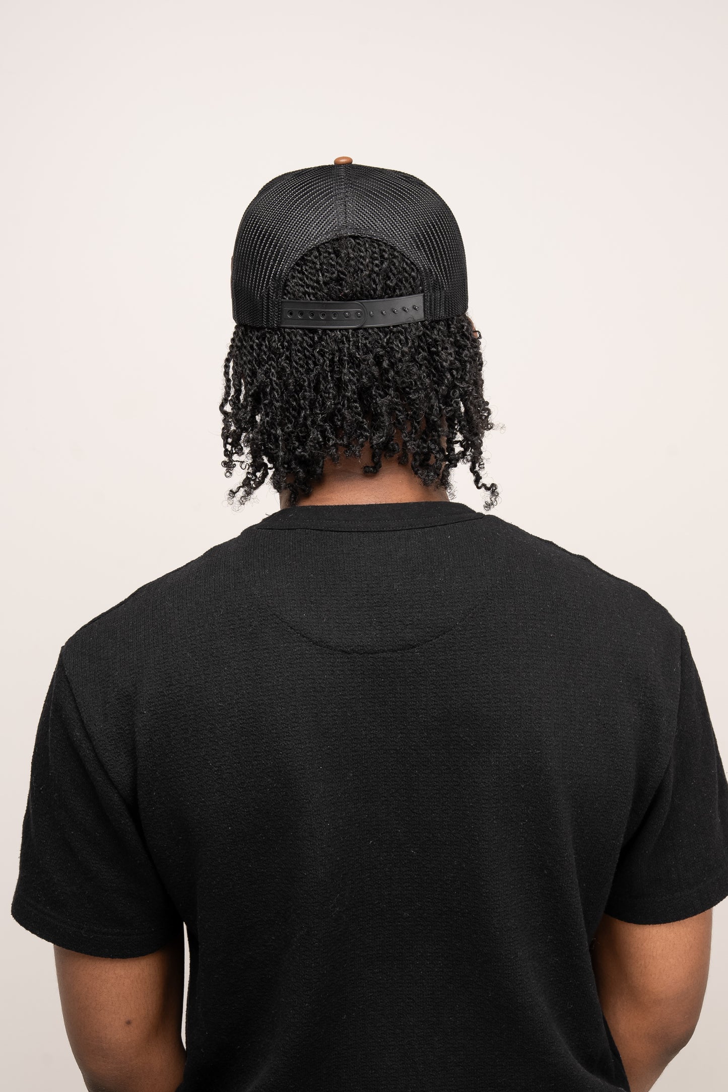 Person facing away from the camera, wearing a black t-shirt and a Guinness Brown Leather Trucker Cap from Guinness Webstore UK, with curly hair visible beneath the mesh back.