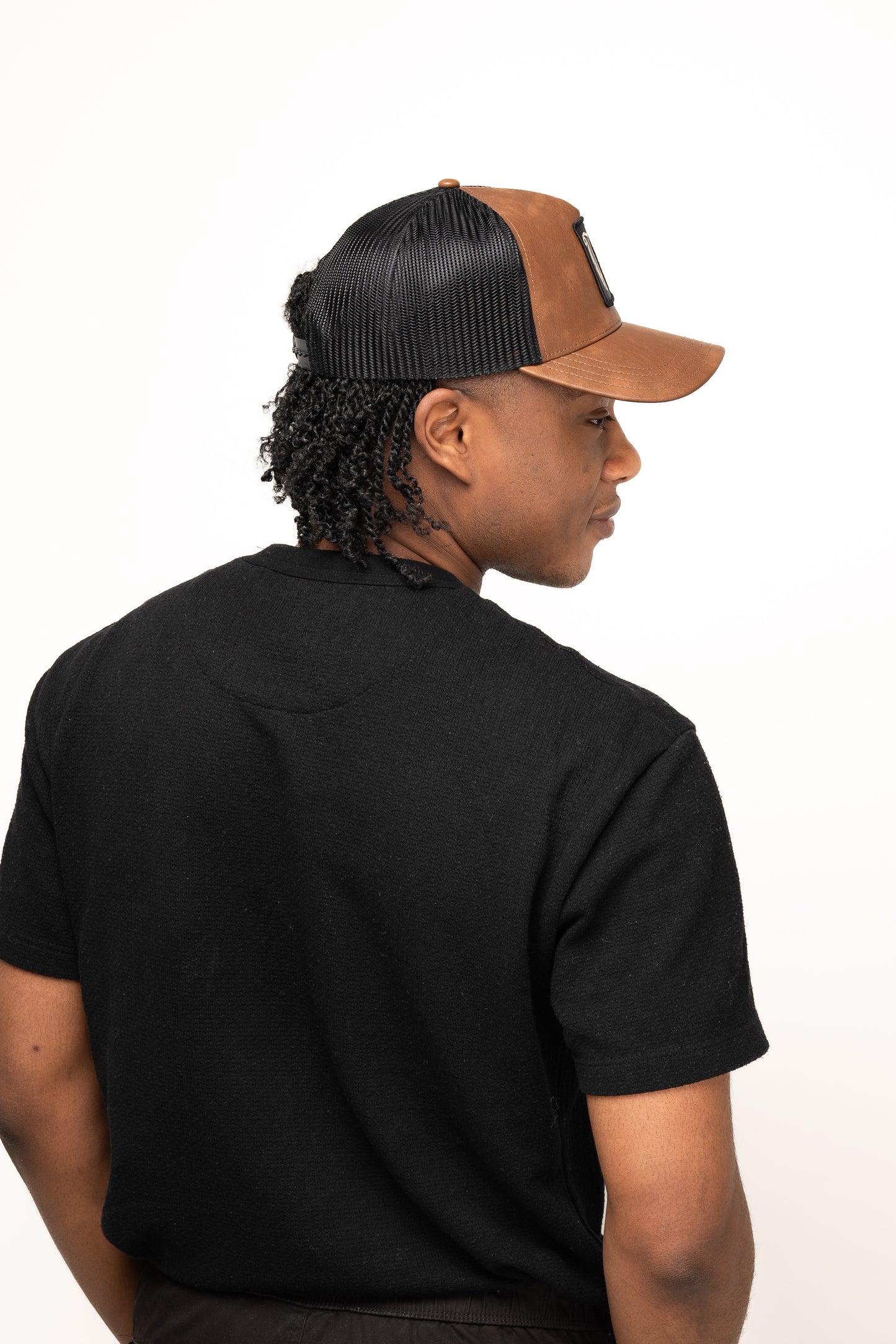 A person in a black t-shirt and a Guinness Brown Leather Trucker Cap from Guinness Webstore UK, featuring the Guinness harp logo, is facing away and looking over their shoulder against a plain light background.