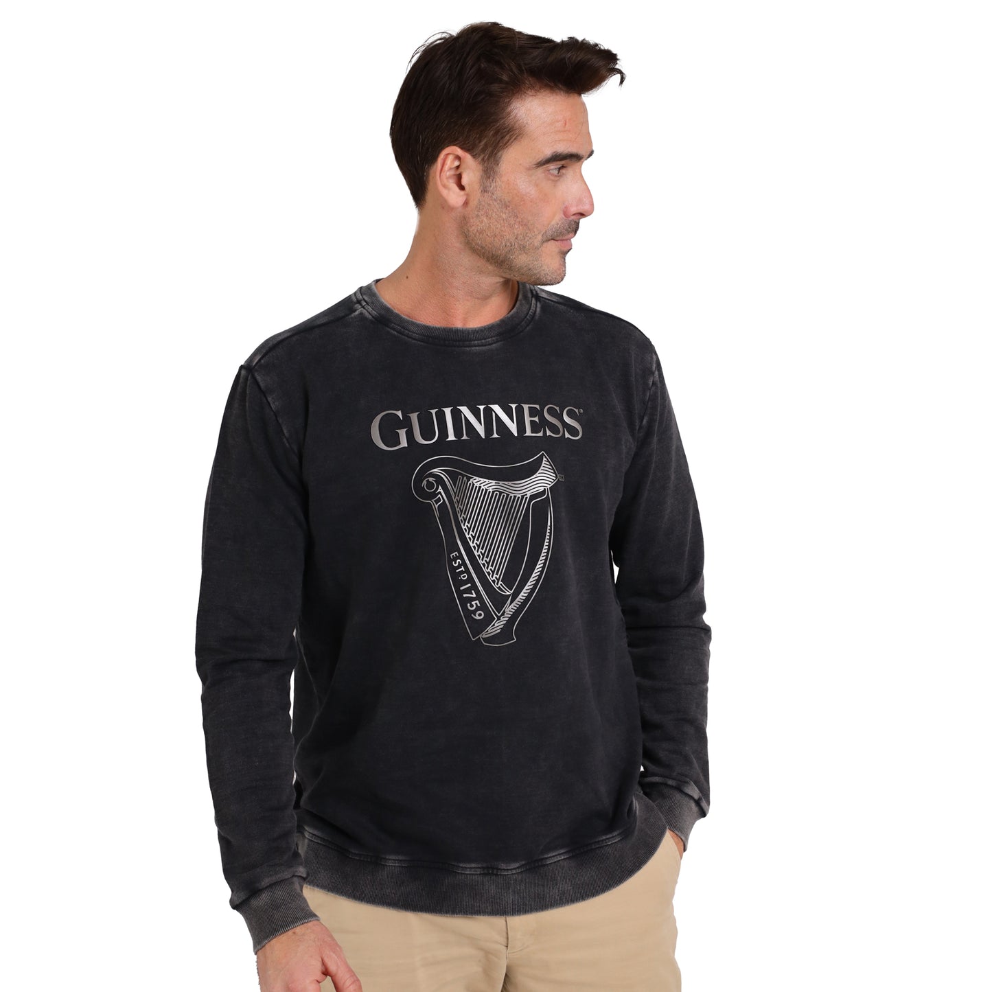 A man in a black Guinness Metallic Print Trademark Sweatshirt from Guinness Webstore UK, made of sustainable cotton and featuring a metallic harp logo, looks to his left with one hand in his pocket.