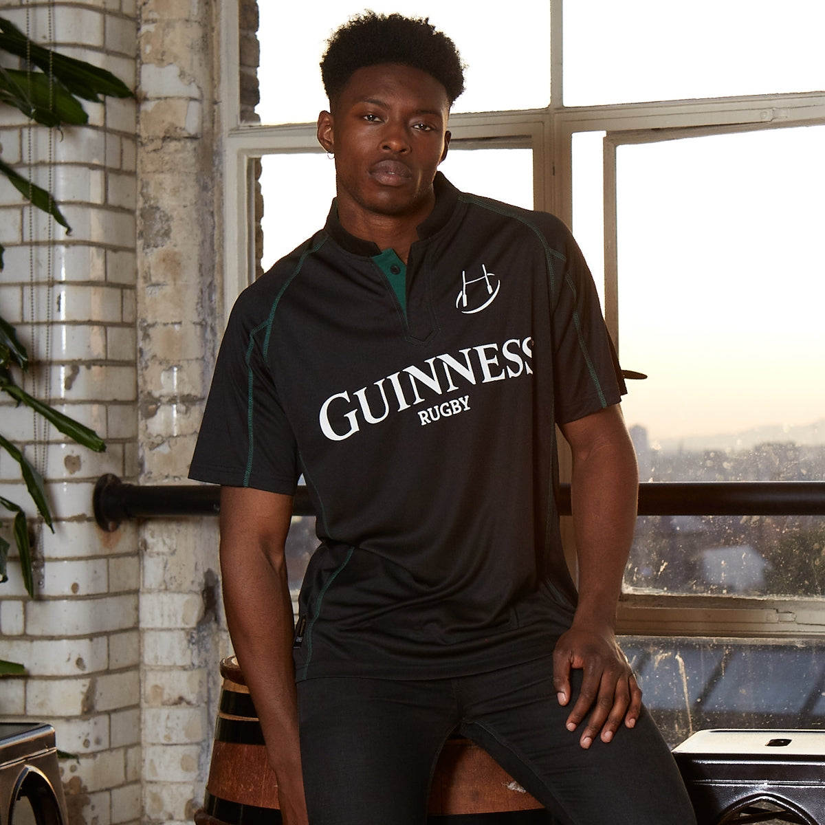A man wearing the Guinness Black & Green Short Sleeve Rugby Jersey sits on a barrel near a large window, surrounded by lush plants and white brick walls.