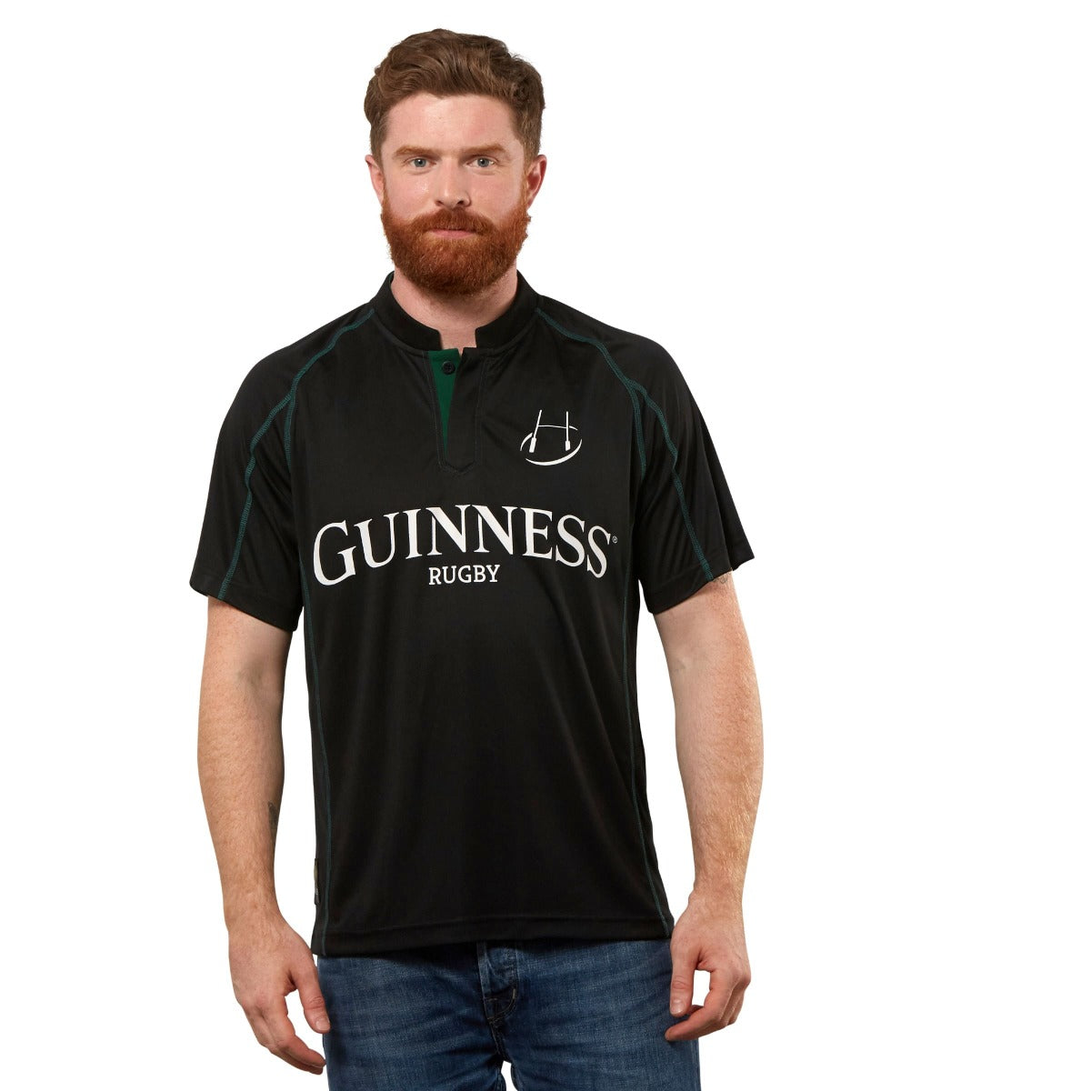 A bearded man stands facing forward, wearing the Guinness Black & Green Short Sleeve Rugby Jersey and blue jeans against a plain white background.