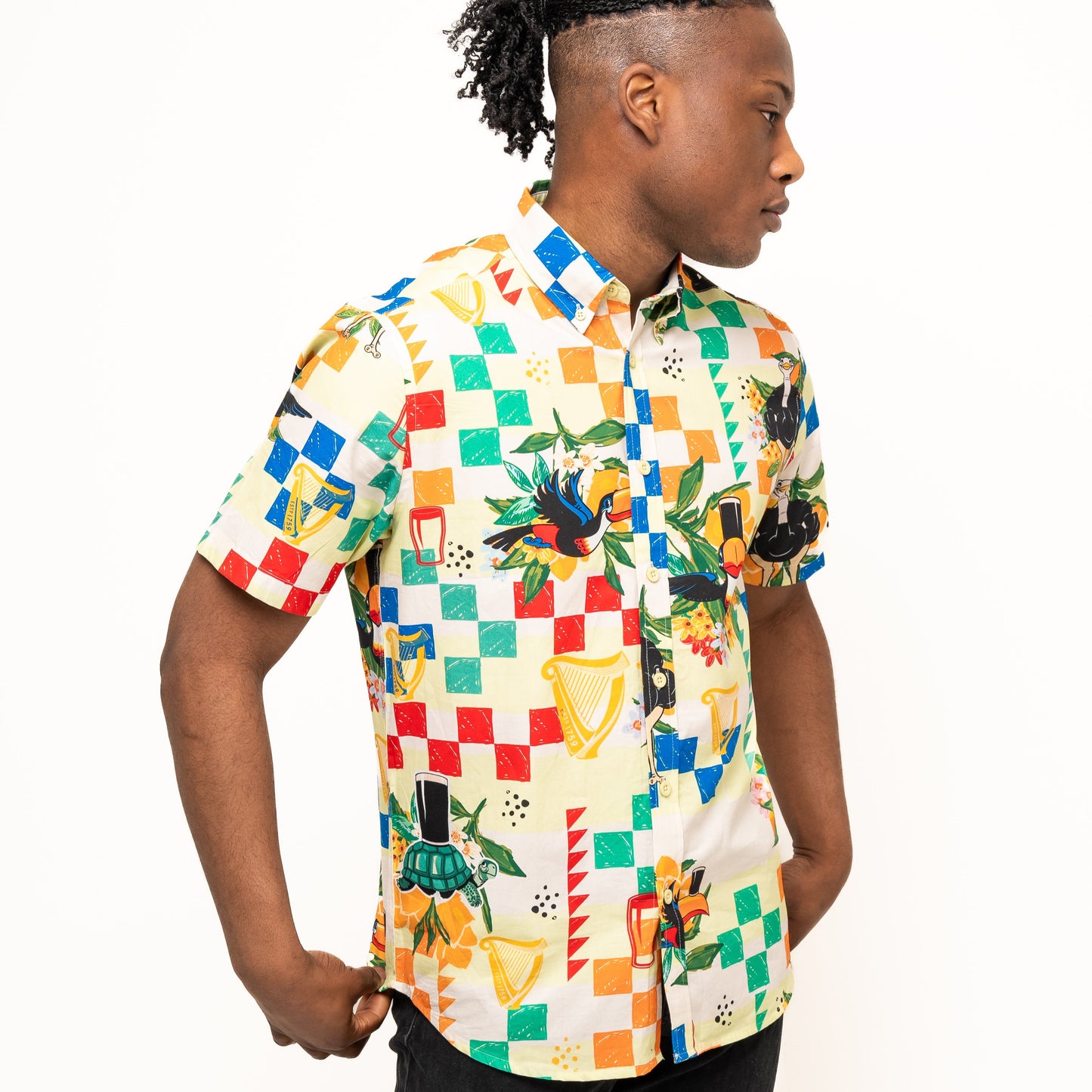 A person wears the Guinness Toucan Beach Shirt by Guinness Webstore UK, a colorful Hawaiian-style short-sleeve cotton button-up, standing against a plain white background facing right with hands in pockets.