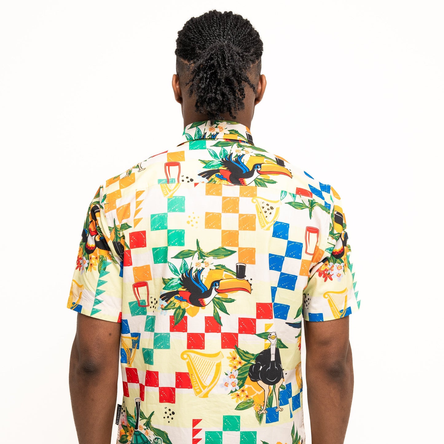 A person with braided hair is seen from behind, wearing the Guinness Toucan Beach Shirt by Guinness Webstore UK—a Hawaiian-style cotton button-up featuring vibrant abstract patterns with birds, flowers, and geometric shapes.