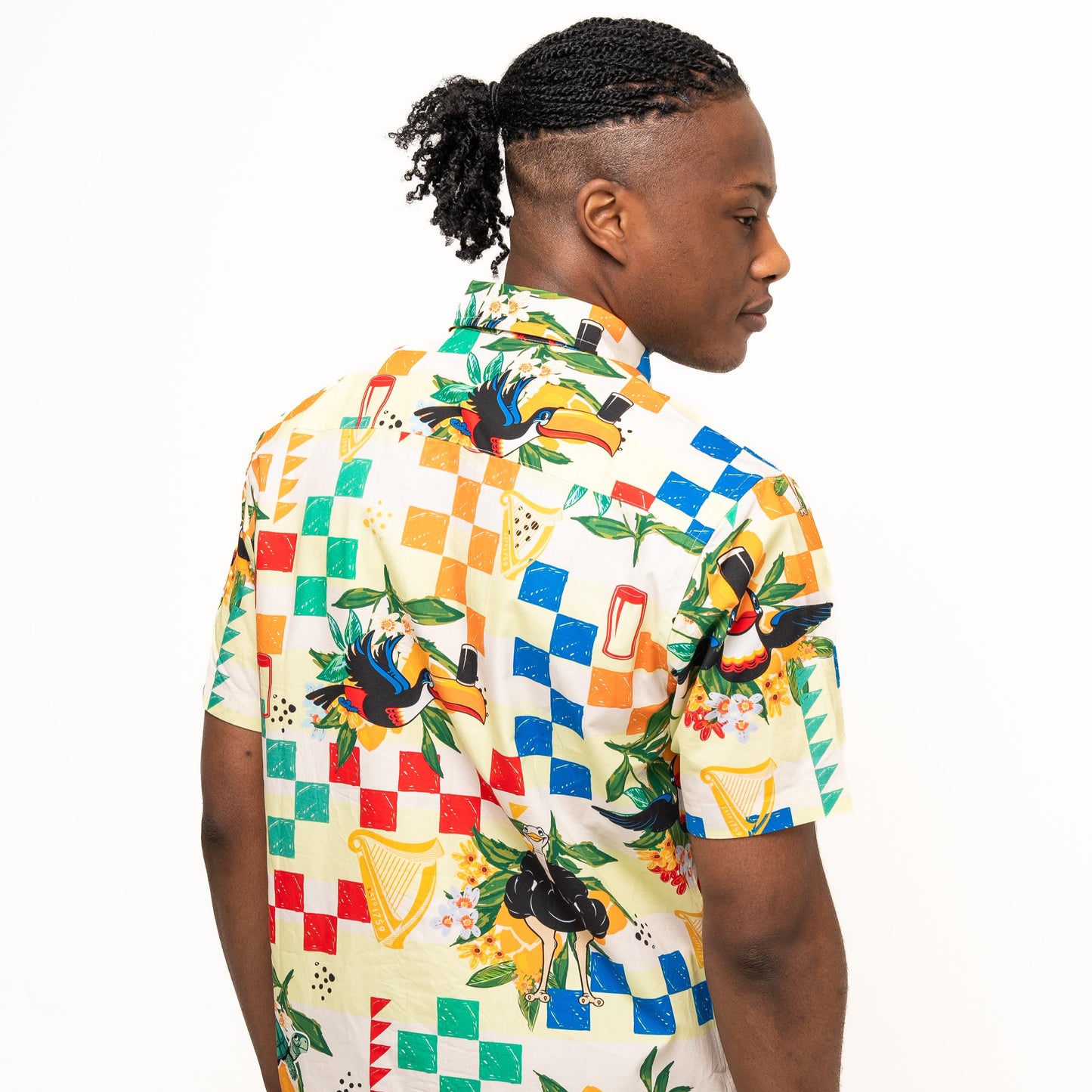 A man with braided hair in a ponytail models the Guinness Toucan Beach Shirt from Guinness Webstore UK, featuring a colorful Hawaiian design in breathable woven cotton, against a white background, facing slightly away from the camera.
