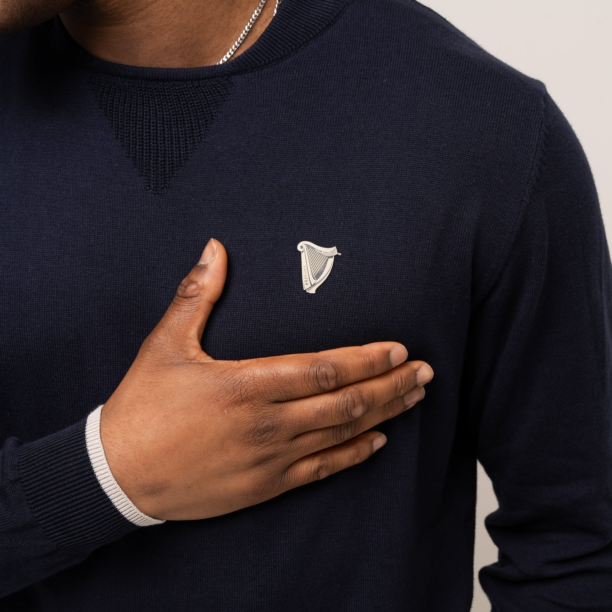 Wearing the navy Guinness Performance Cotton Sweater from Guinness Webstore UK, a person places their right hand over their chest, highlighting its moisture-wicking comfort—ideal for a day on the golf course.