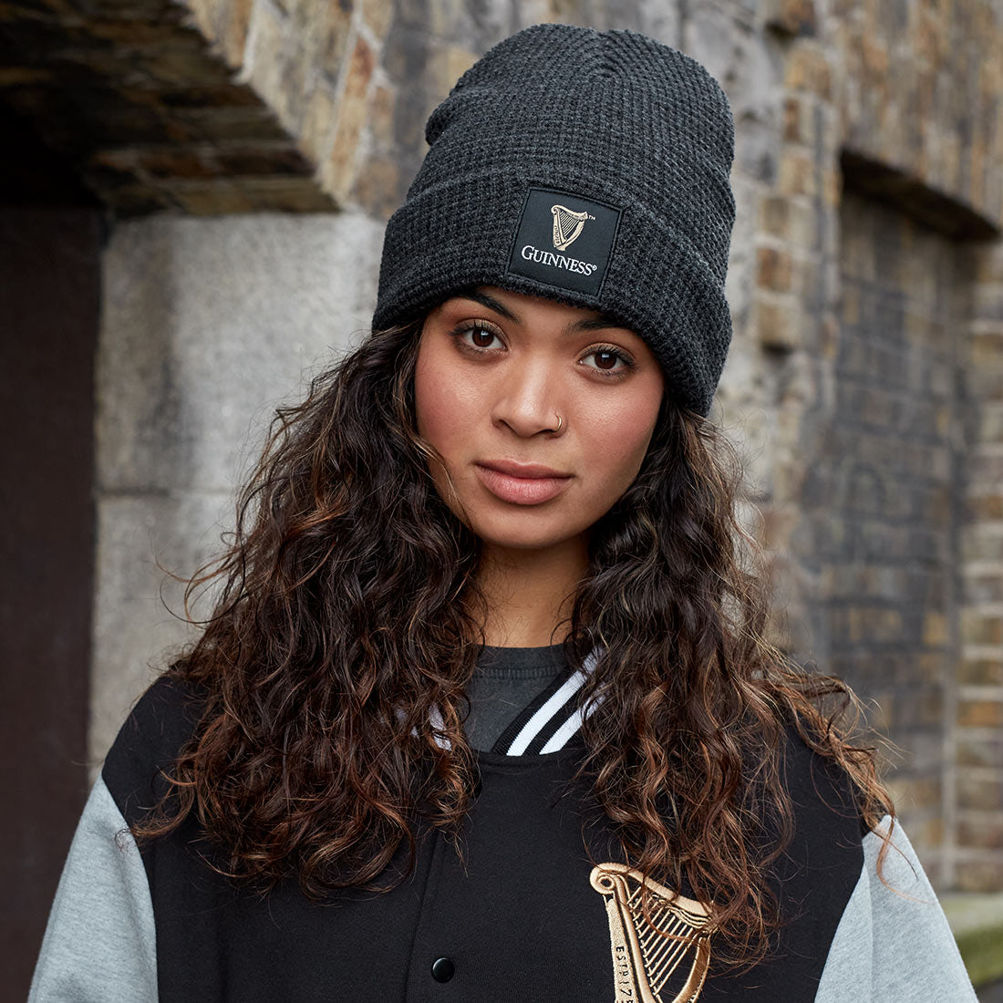 A woman with curly hair wears a Guinness UK Thermal Warm Beanie and a black jacket with a gold harp emblem, standing in front of a stone wall.