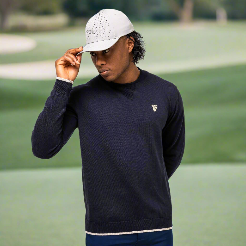 A man in a Guinness Performance Cotton Sweater - Navy from Guinness Webstore UK and a light cap stands on a golf course, holding the brim of his cap with one hand.