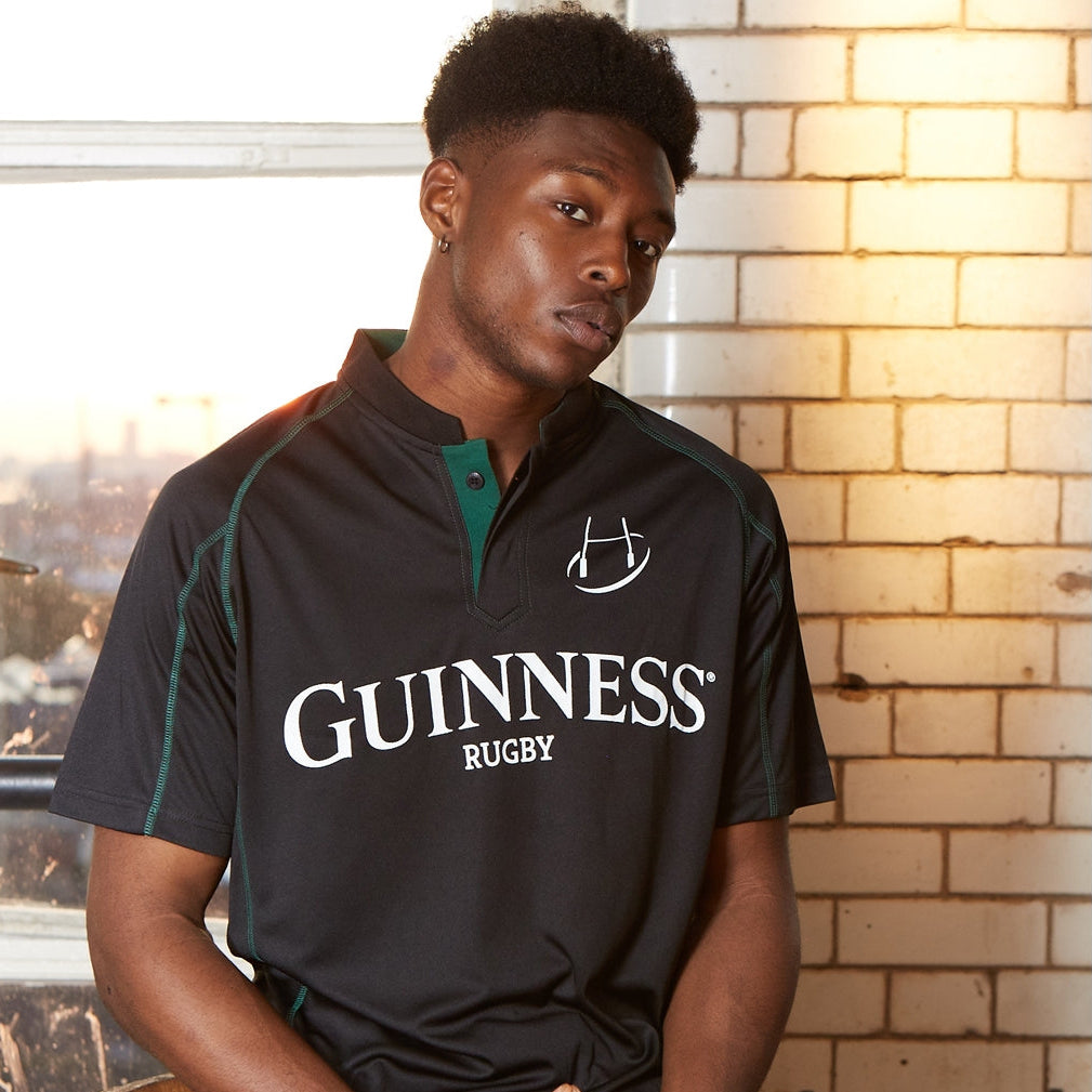 A man wearing a Guinness Black & Green Short Sleeve Rugby Jersey sits indoors by a window and white tiled wall, illuminated by natural light.