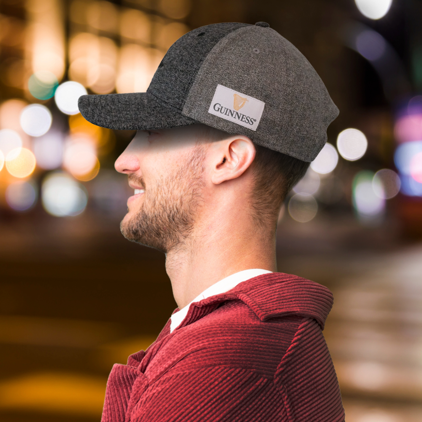 A man in a red zip-up shirt and a Guinness Heathered Wool Grey Cap from Guinness Webstore UK stands in profile against a blurred city street background at night.