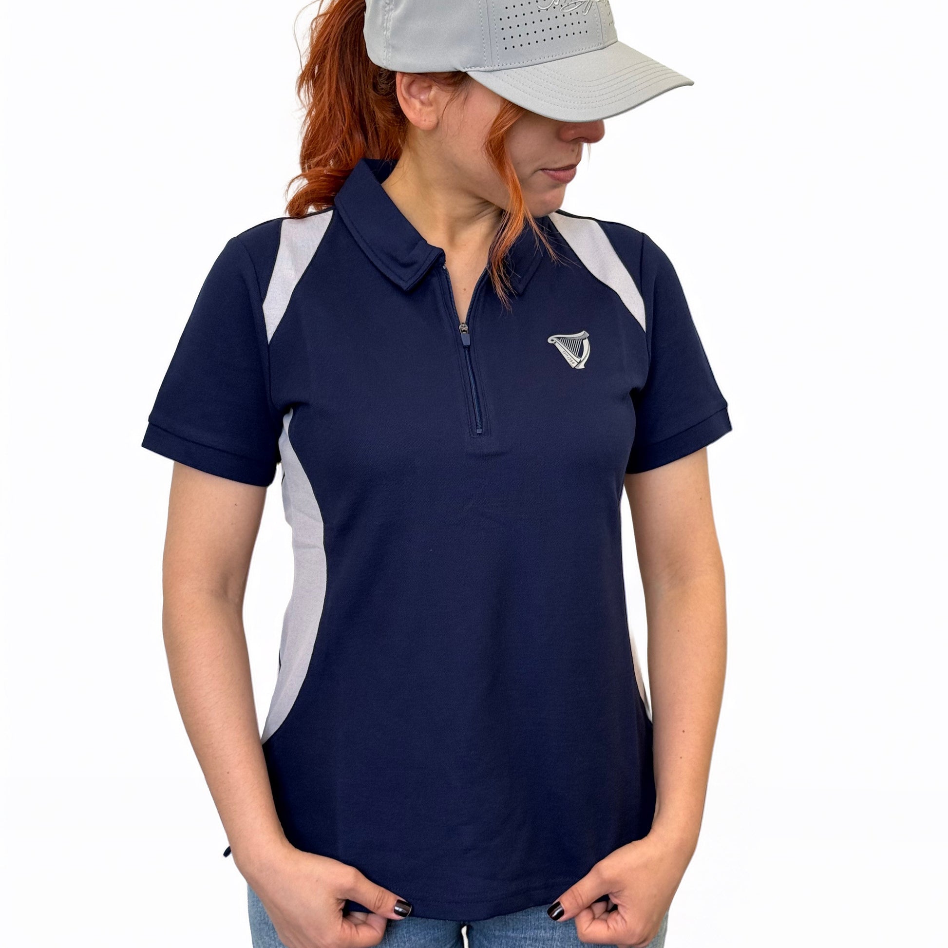 A woman wearing a blue Guinness Webstore UK Ladies Performance Polo and a gray cap stands with her hands in her pockets, looking to the side against a plain white background.