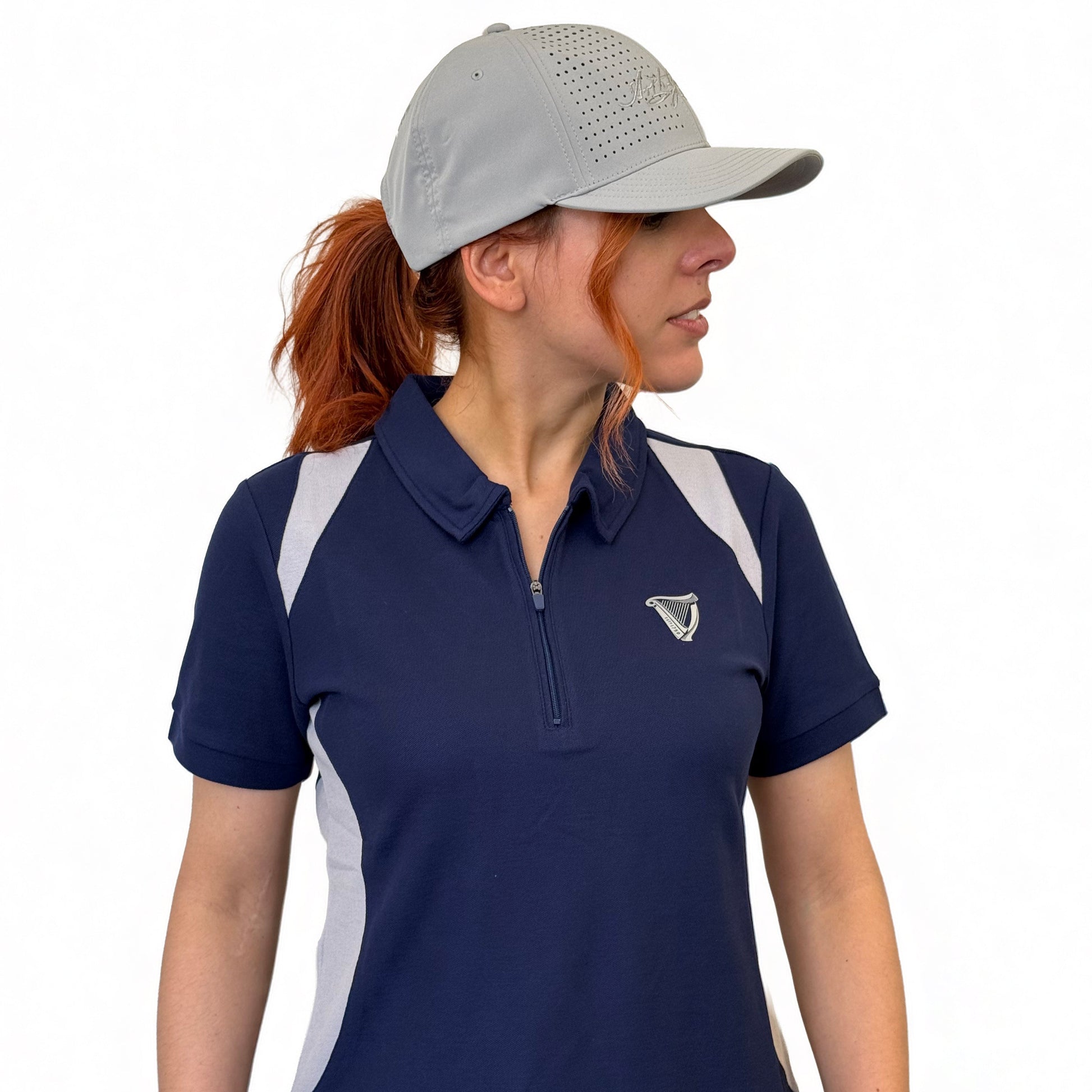 A woman with reddish hair in a ponytail, wearing a gray cap and the Ladies Performance Polo - Blue from Guinness Webstore UK, looks to the side against a plain white background.
