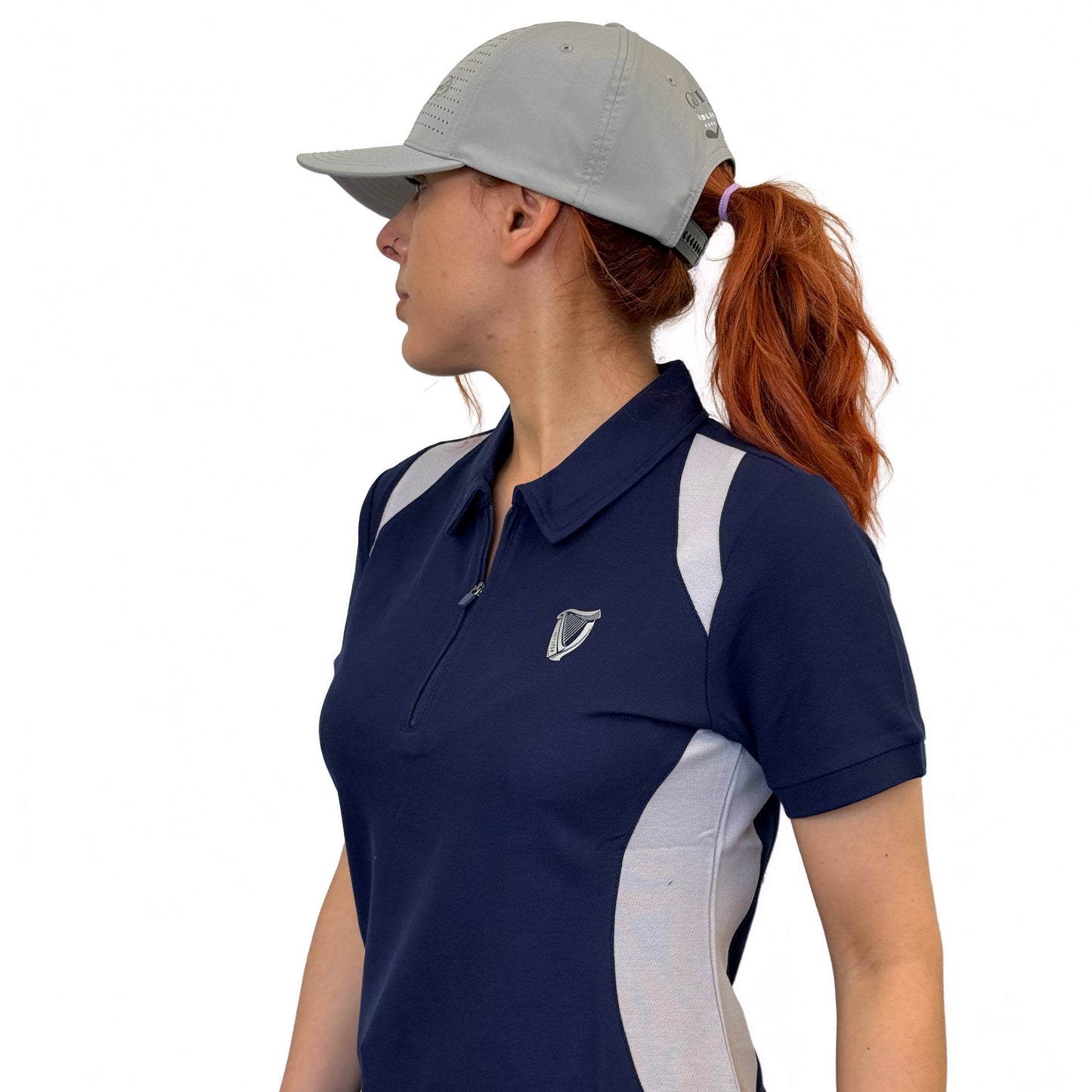 A woman with long red hair in a ponytail wears a gray cap and the Guinness Webstore UK Ladies Performance Polo - Blue, featuring white side panels, posing against a plain white background.
