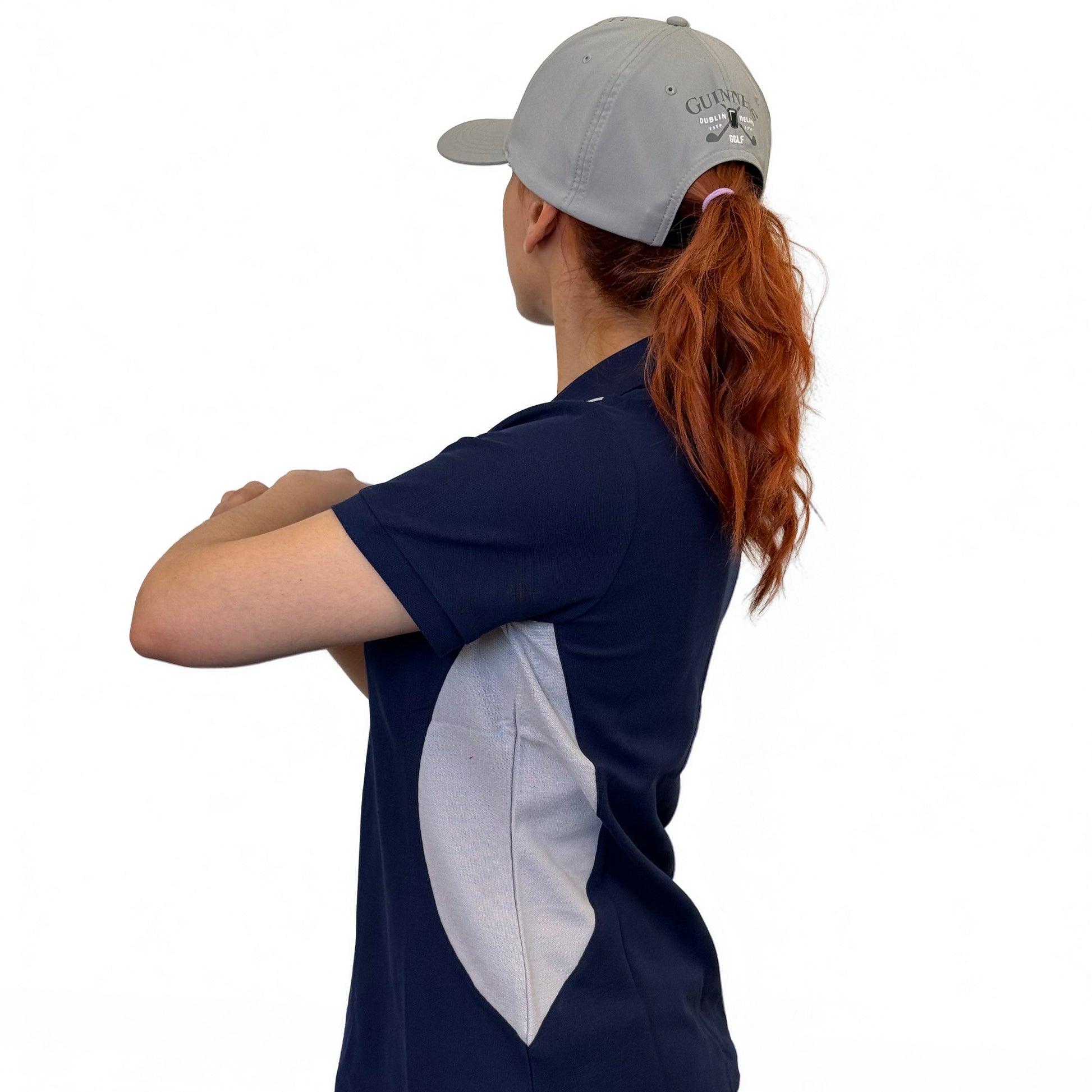 A woman with long red hair wears a gray cap and the Guinness Webstore UK Ladies Performance Polo in blue, made from premium double pique fabric, standing with arms crossed against a white background—perfect for the golf course.