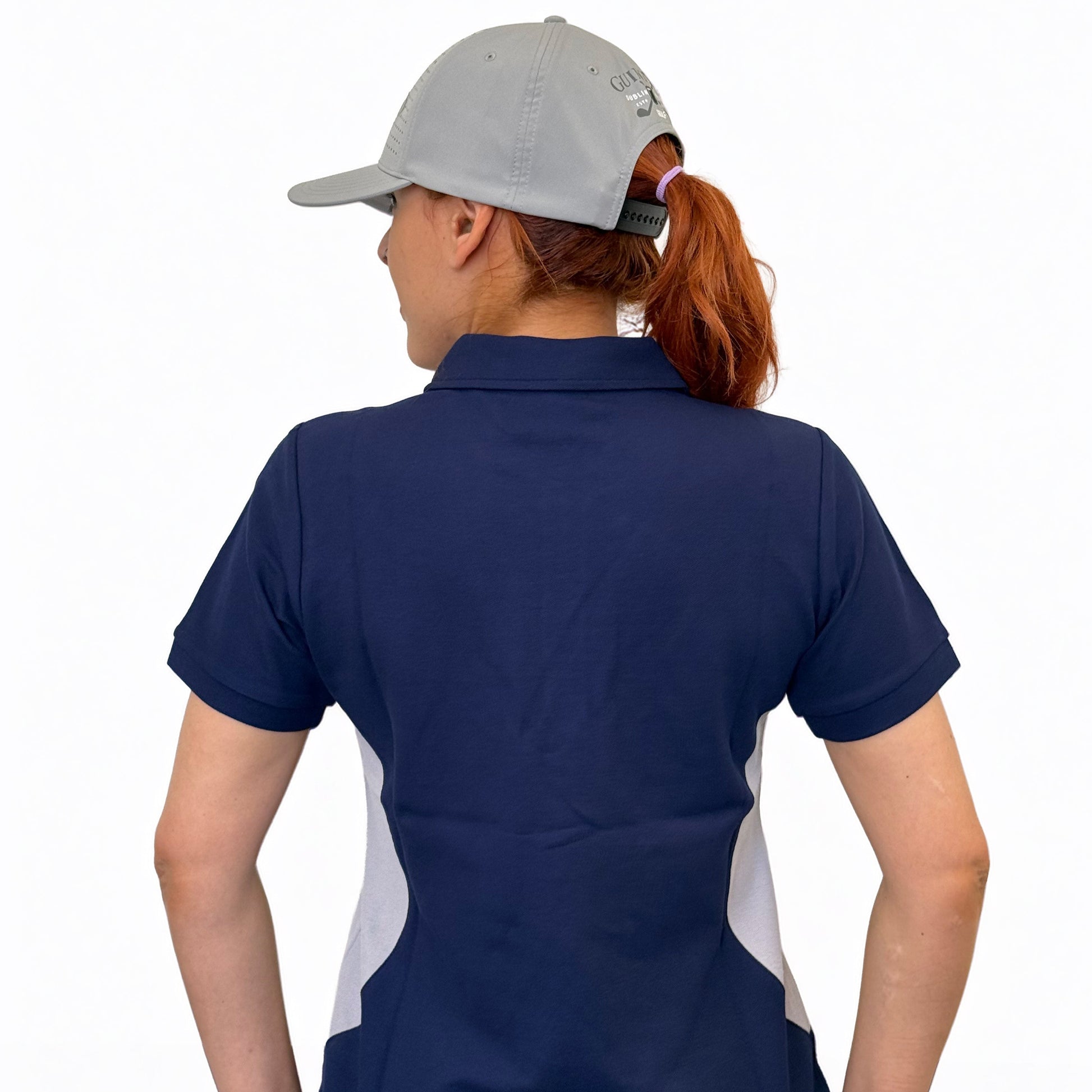 A person with red hair in a ponytail wears a light gray cap and a Guinness Webstore UK Ladies Performance Polo - Blue, made from premium double pique fabric, standing with their back to the camera on a golf course.