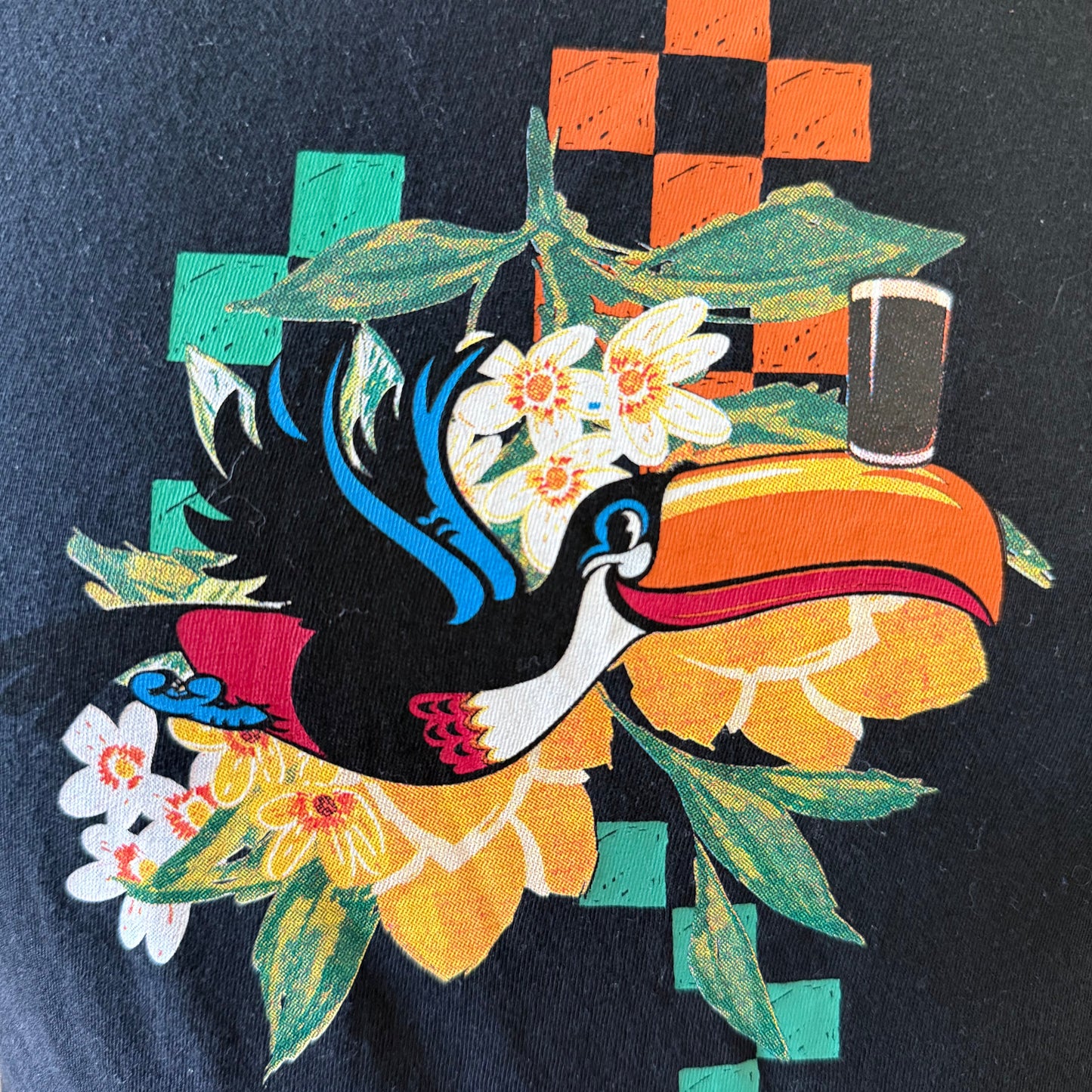 The Guinness Toucan Ruched Ladies Black Top from Guinness Webstore UK features a vibrant toucan with an orange beak, floral accents, and bold geometric shapes—an eye-catching piece inspired by Irish culture.