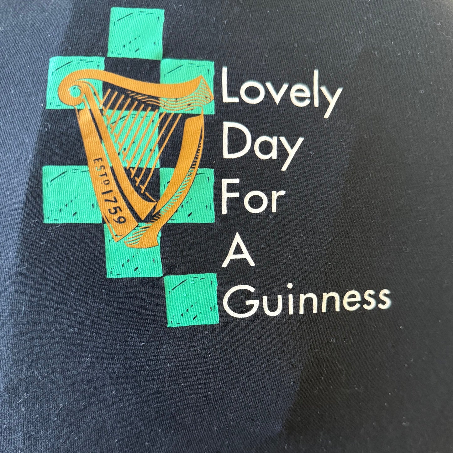 The Guinness Toucan Ruched Ladies Black Top from Guinness Webstore UK features a gold harp and green cross design with the phrase "Lovely Day For A Guinness" on a dark background, celebrating Irish culture.
