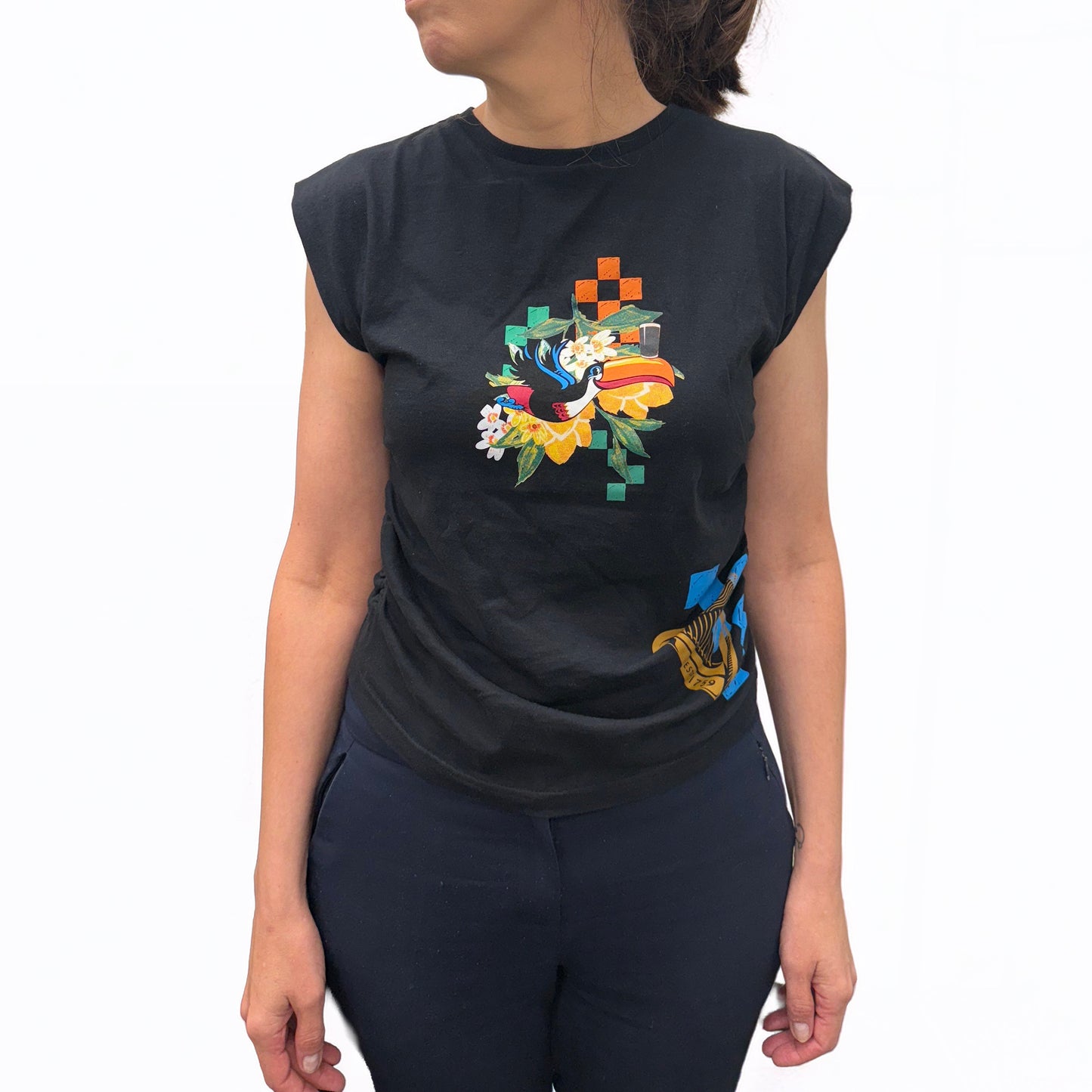 A woman wears the Guinness Toucan Ruched Ladies Black Top from Guinness Webstore UK, featuring a vibrant abstract floral and geometric design on the front, paired with dark pants against a white background.
