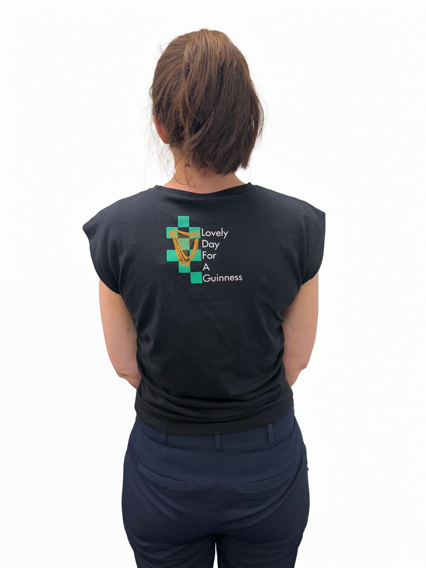 A person stands with their back to the camera wearing the Guinness Toucan Ruched Ladies Black Top from the Guinness Webstore UK, featuring a pint graphic and Irish-inspired "Lovely Day For A Guinness" slogan.
