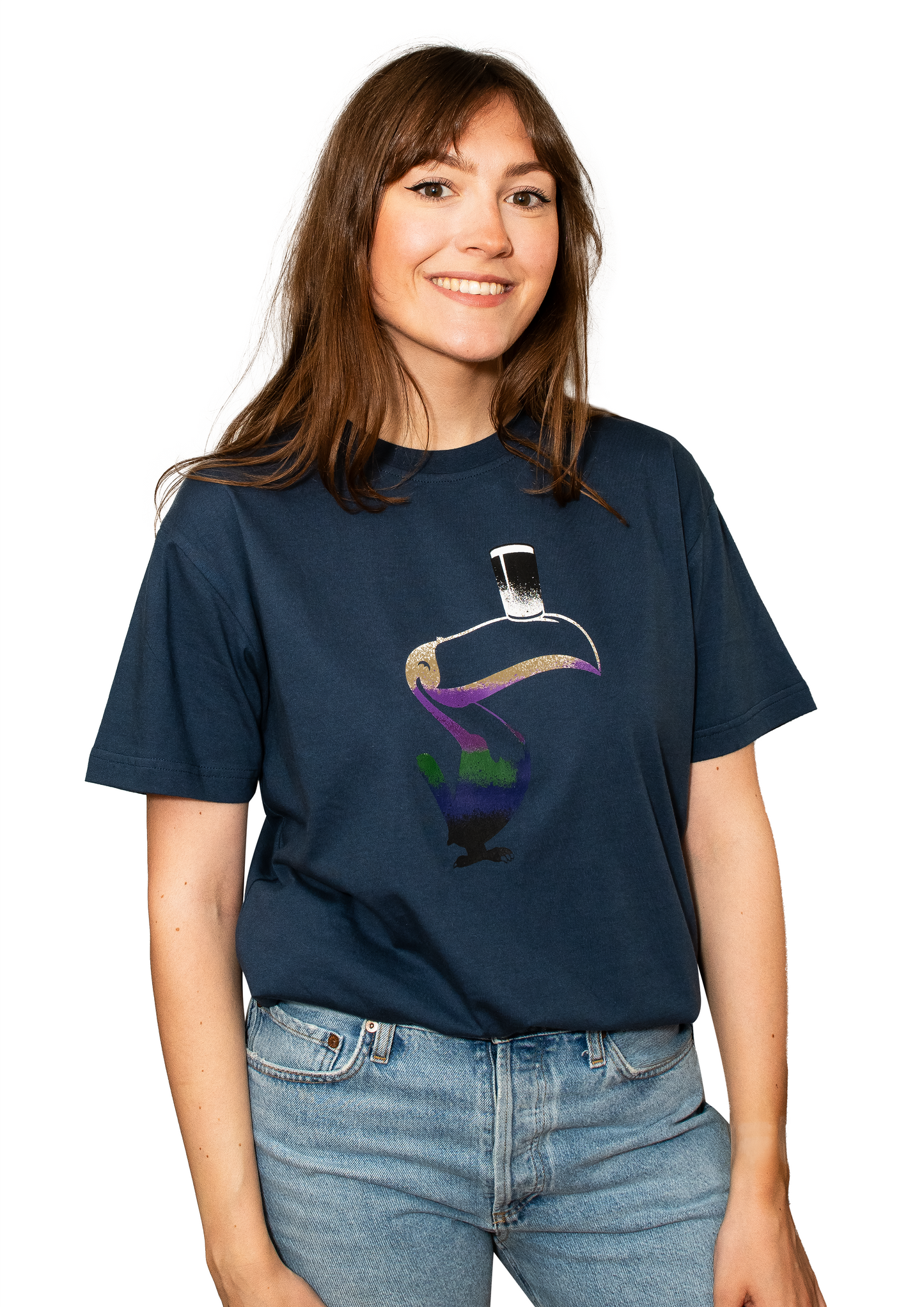 A woman in light blue jeans stands smiling, wearing a navy Guinness Liquid Toucan T-Shirt made from sustainable BCI cotton and featuring a vibrant toucan graphic.
