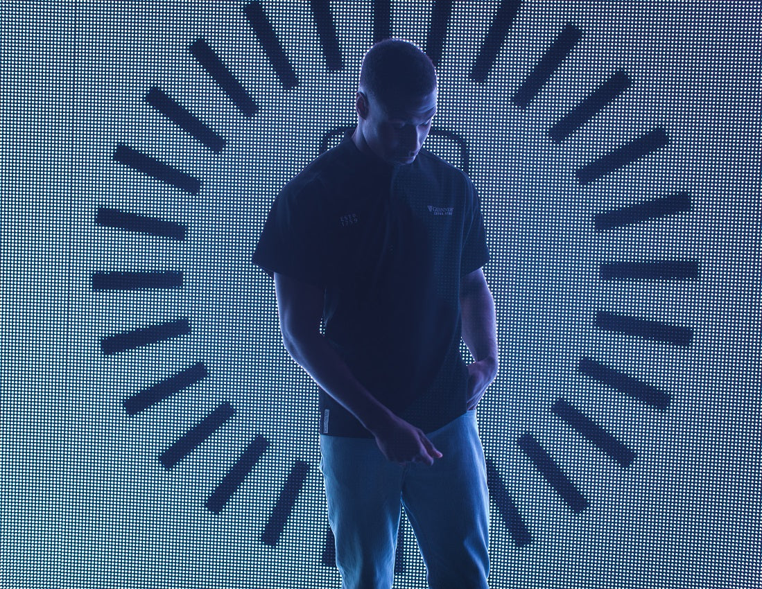 A man in a black shirt standing in front of a circular light with Guinness UK branding and wearing the All Black Rugby Jersey.