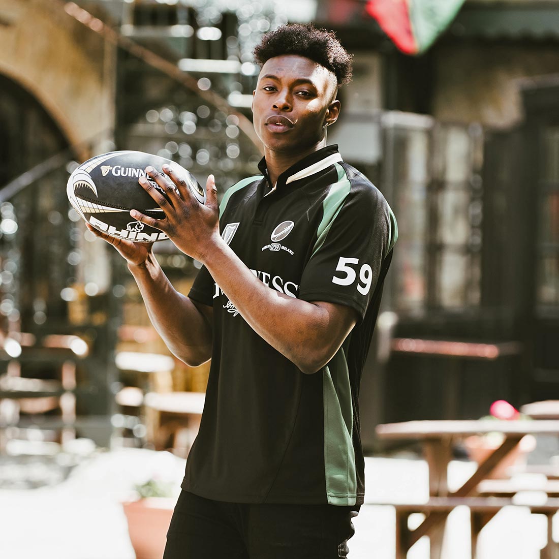 A young man, wearing sportswear, holding a Guinness Black and Green Short sleeve performance Rugby Jersey in front of a restaurant.