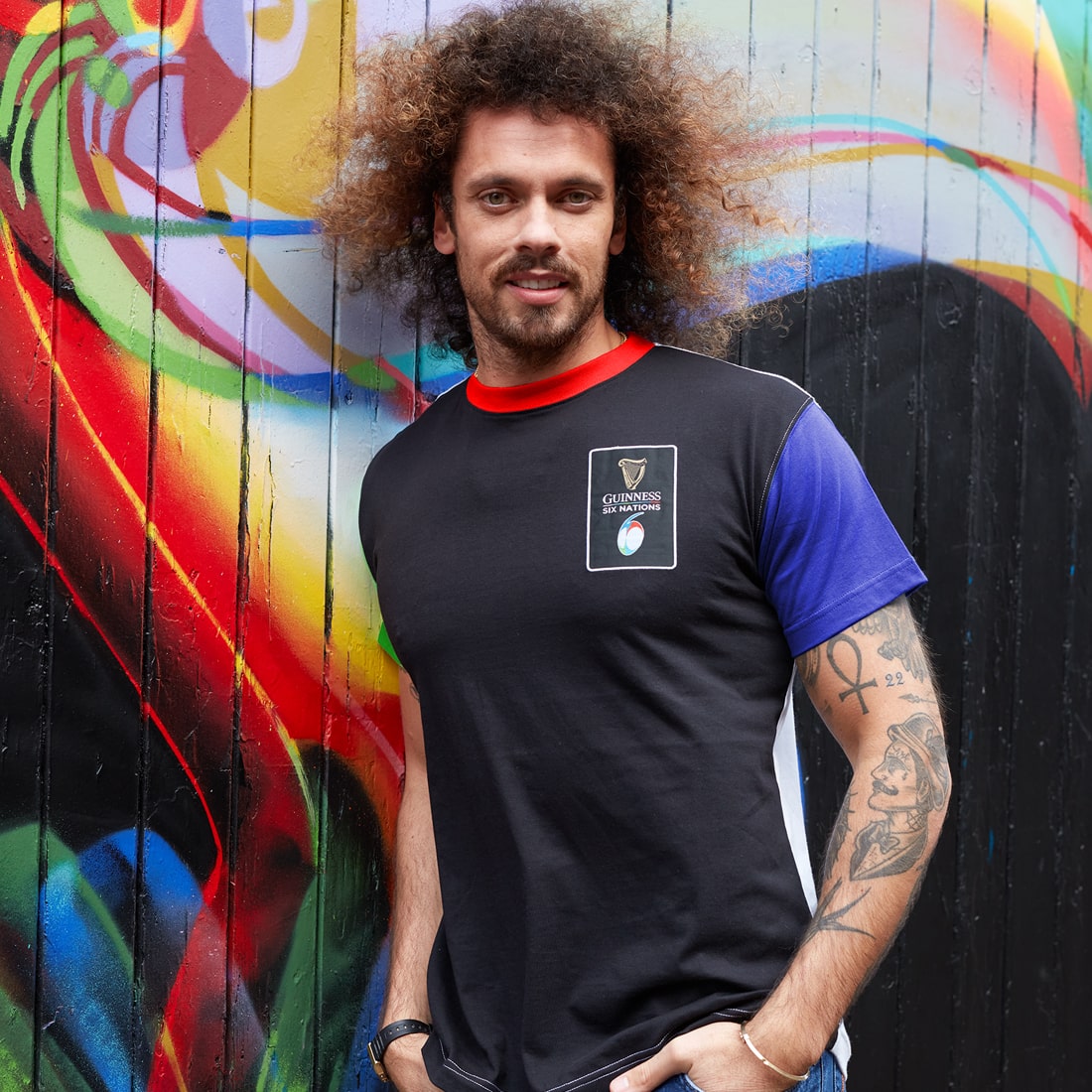 A man with curly hair wearing a Guinness UK Six Nations Premium Colour Block T-Shirt, standing in front of a graffiti wall.